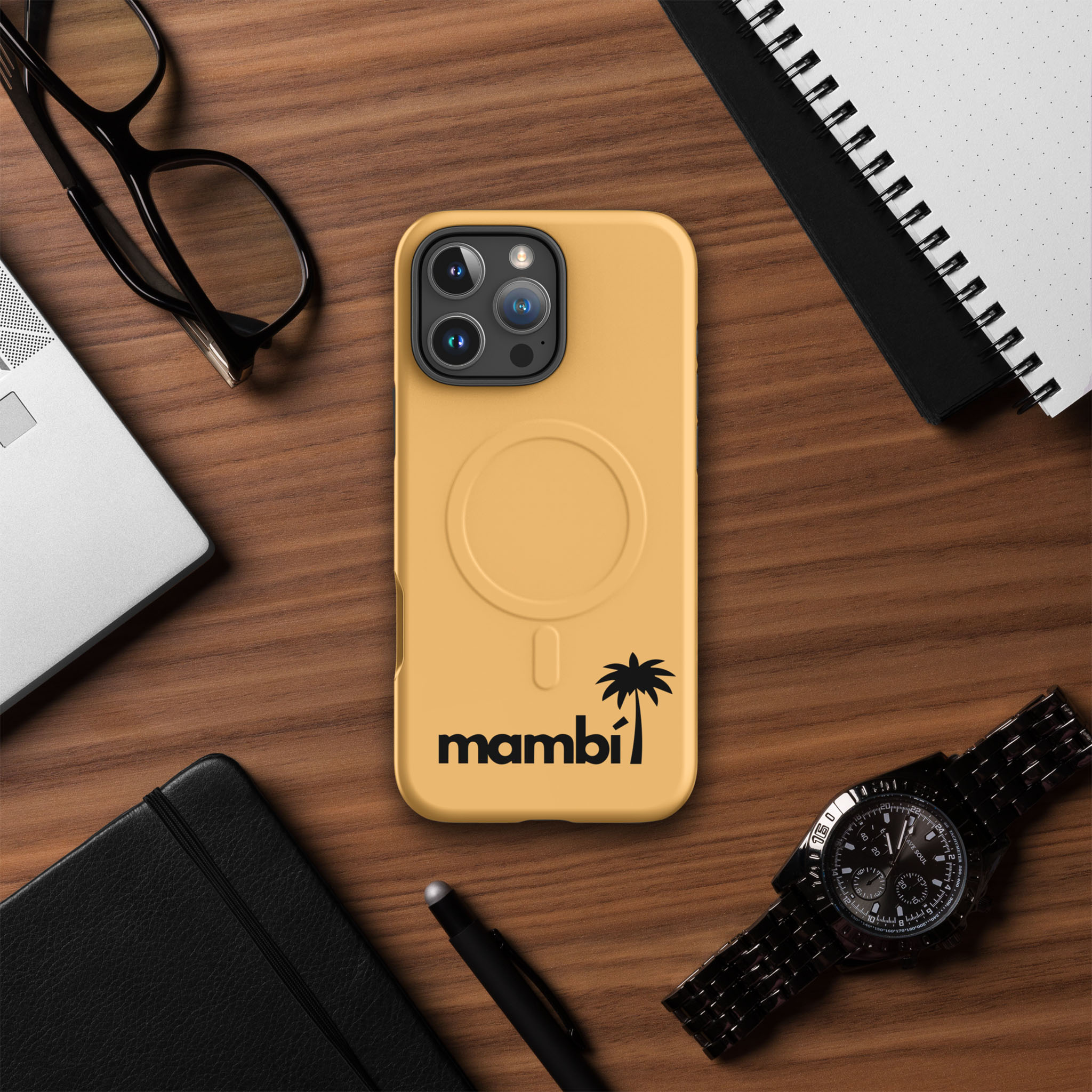 A tan phone with a mambi case featuring a palm tree logo lies on a wooden desk surrounded by eyeglasses, a notebook, a pen, a laptop, and a black wristwatch.