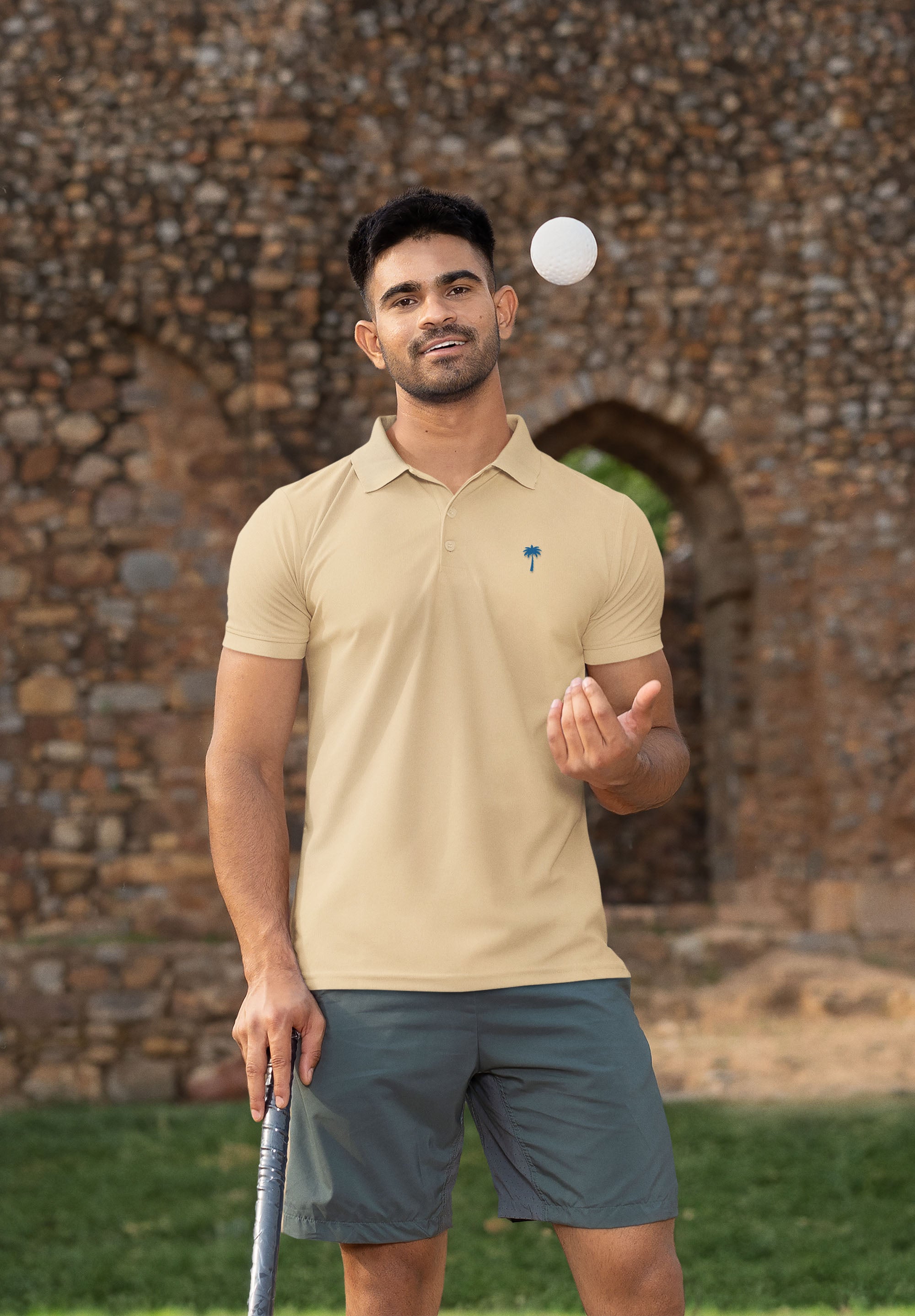 A man in a beige polo shirt and gray shorts stands outdoors, holding a golf club in one hand and tossing a golf ball in the air, with a stone arch and wall in the background.