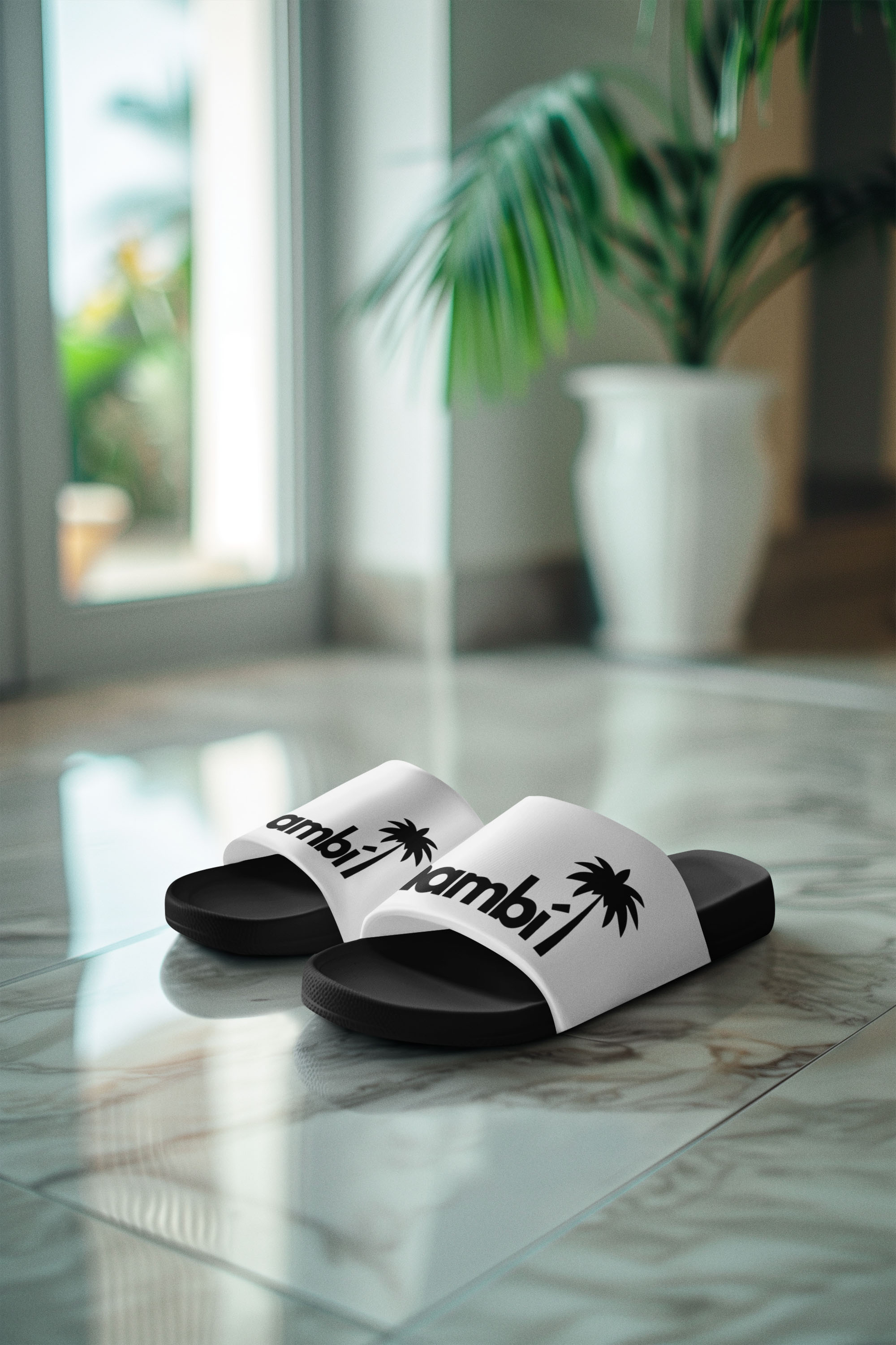 A pair of black-and-white slide sandals with the word bambi and a palm tree graphic on the straps sit on a shiny, tiled floor near a potted plant and glass doors letting in sunlight.