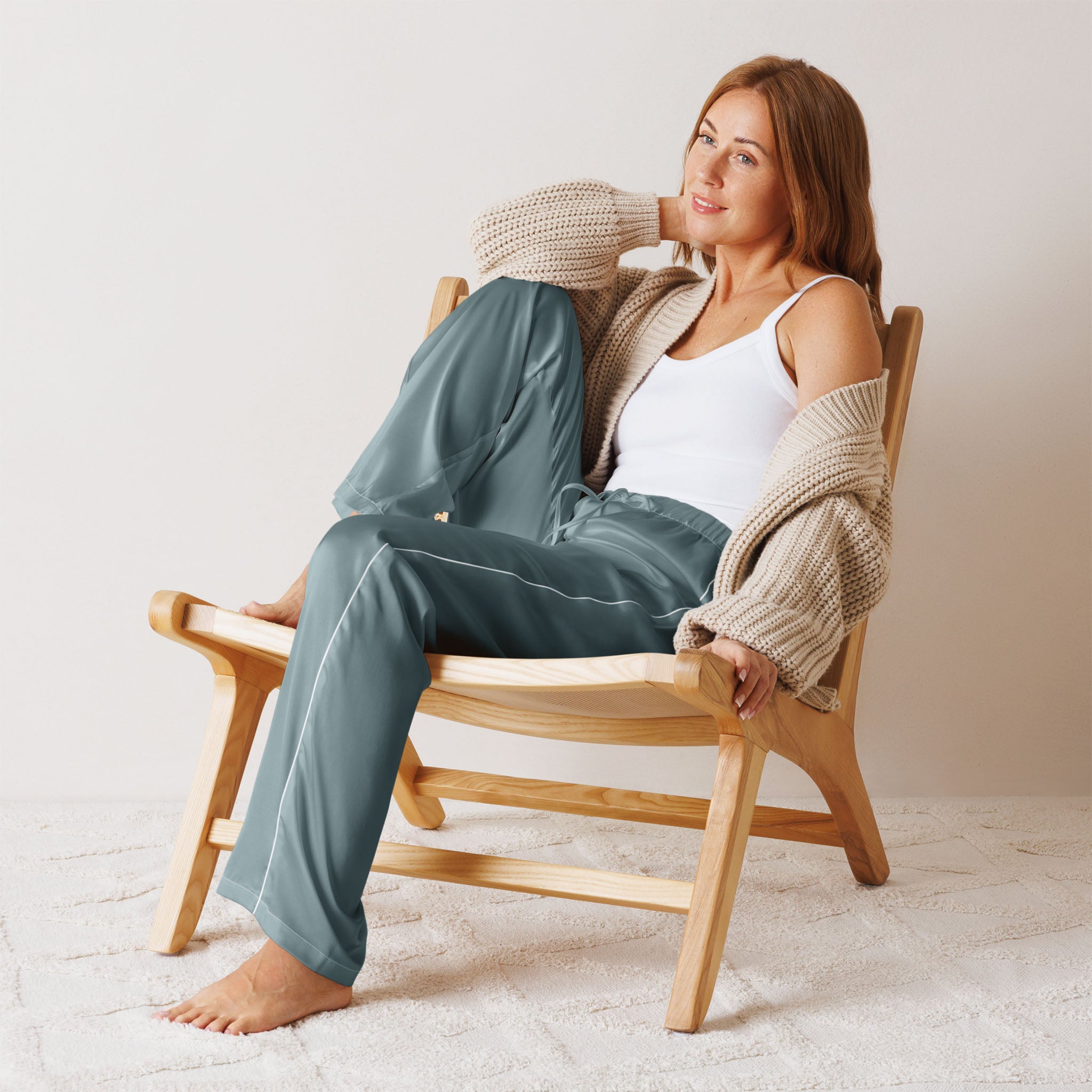 A woman with long brown hair sits relaxed on a wooden chair, wearing a white tank top, blue-green pants, and a chunky beige cardigan. She is barefoot and smiling softly in a cozy, neutral-toned room.