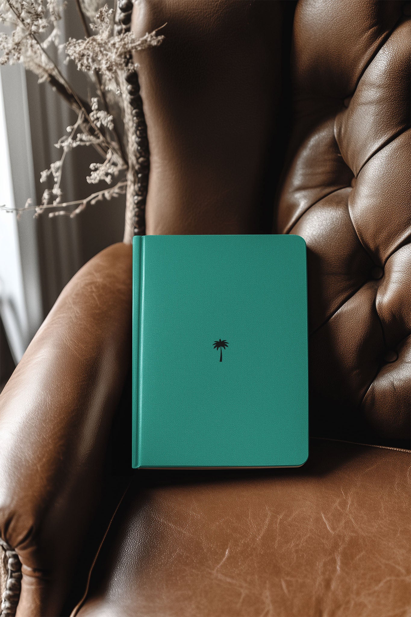 A teal notebook with a small black palm tree design on the cover rests on a tufted brown leather armchair near a window, with dried branches in the background.
