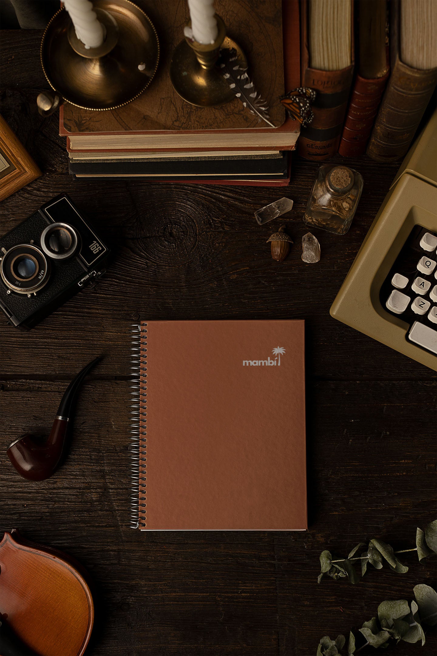 A spiral-bound brown notebook labeled “mambi” is centered on a dark wooden desk, surrounded by vintage items including a camera, violin, tobacco pipe, candlesticks, books, a typewriter, and small decorative objects.