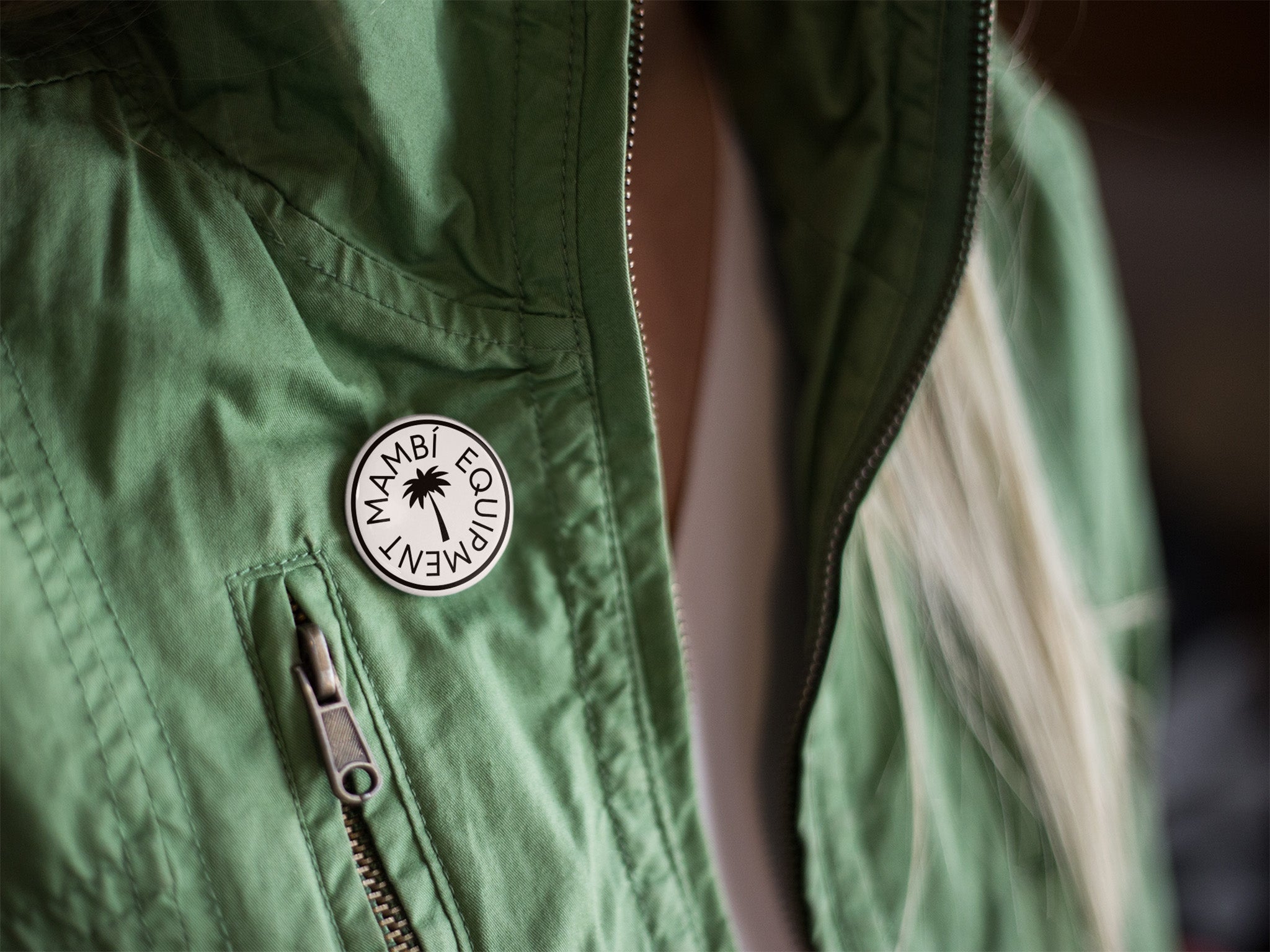 A person wearing a green jacket with a round Mambi Equipment pin featuring a palm tree design attached near the chest zipper.