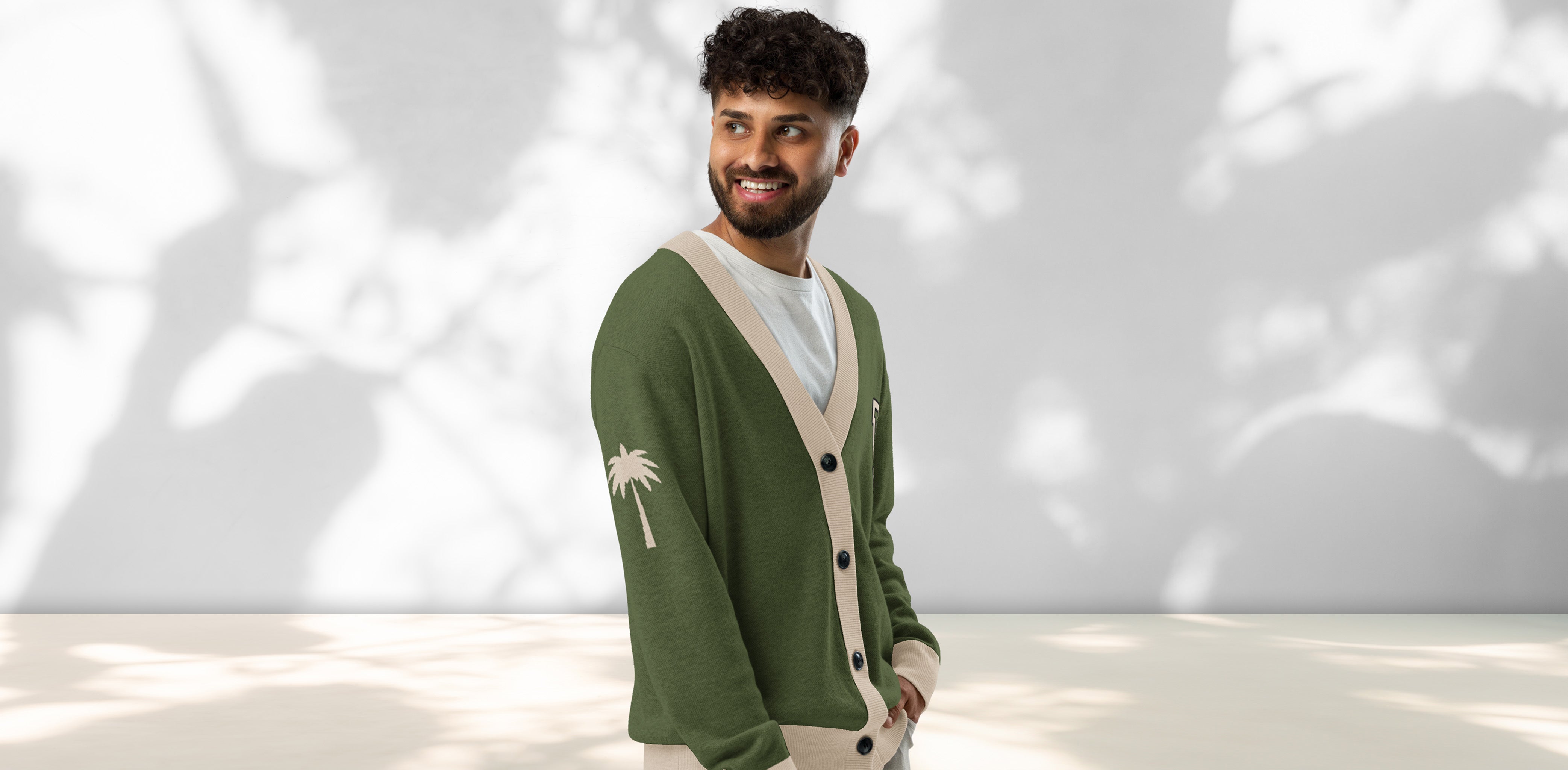 A man with curly hair and a beard, wearing a green and beige cardigan with a palm tree design on the sleeve, stands smiling against a light background with leafy shadows.