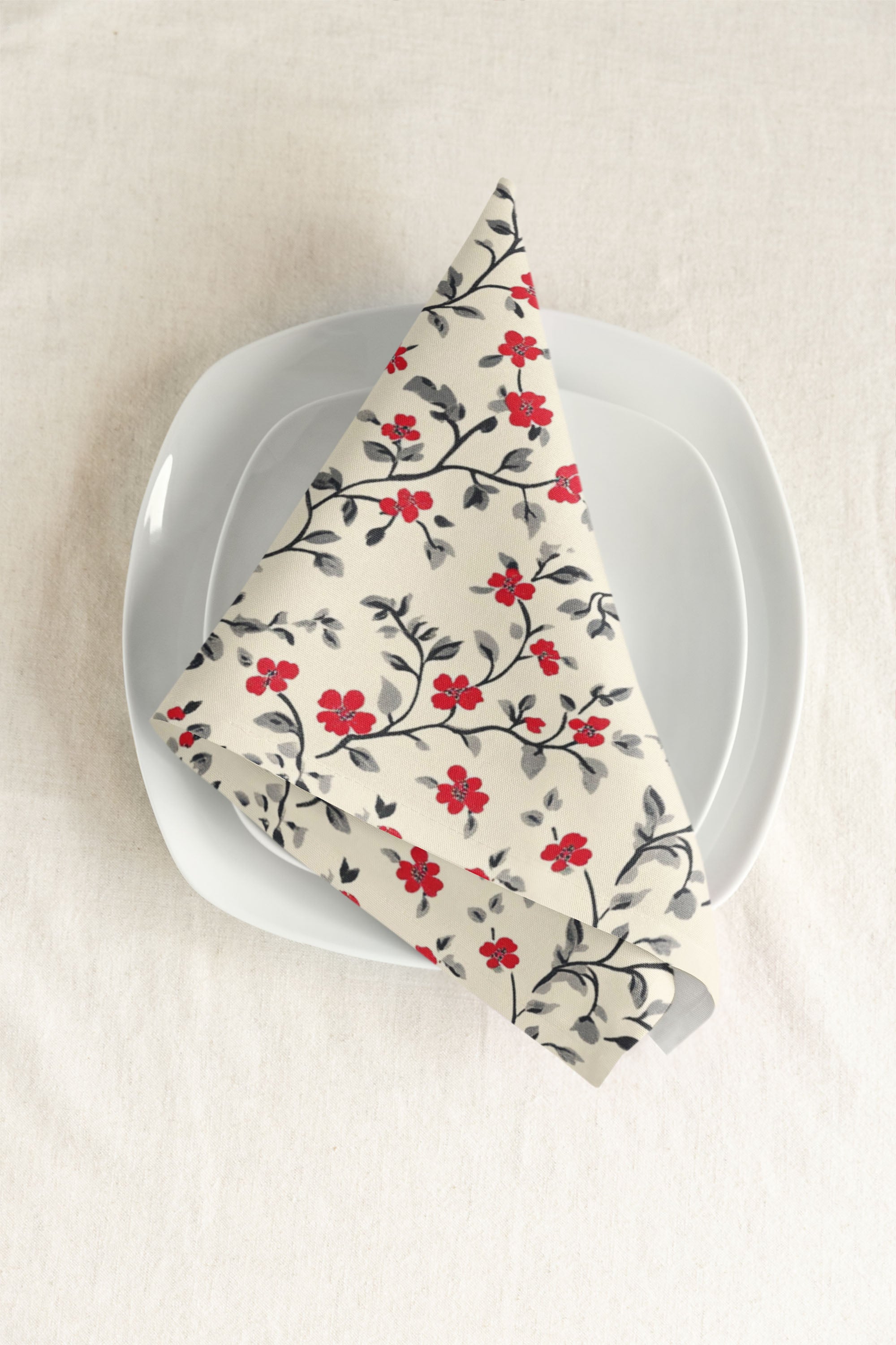 A white plate with a square edge sits on a neutral tablecloth, topped with a neatly folded napkin featuring a pattern of red flowers and black leaves.