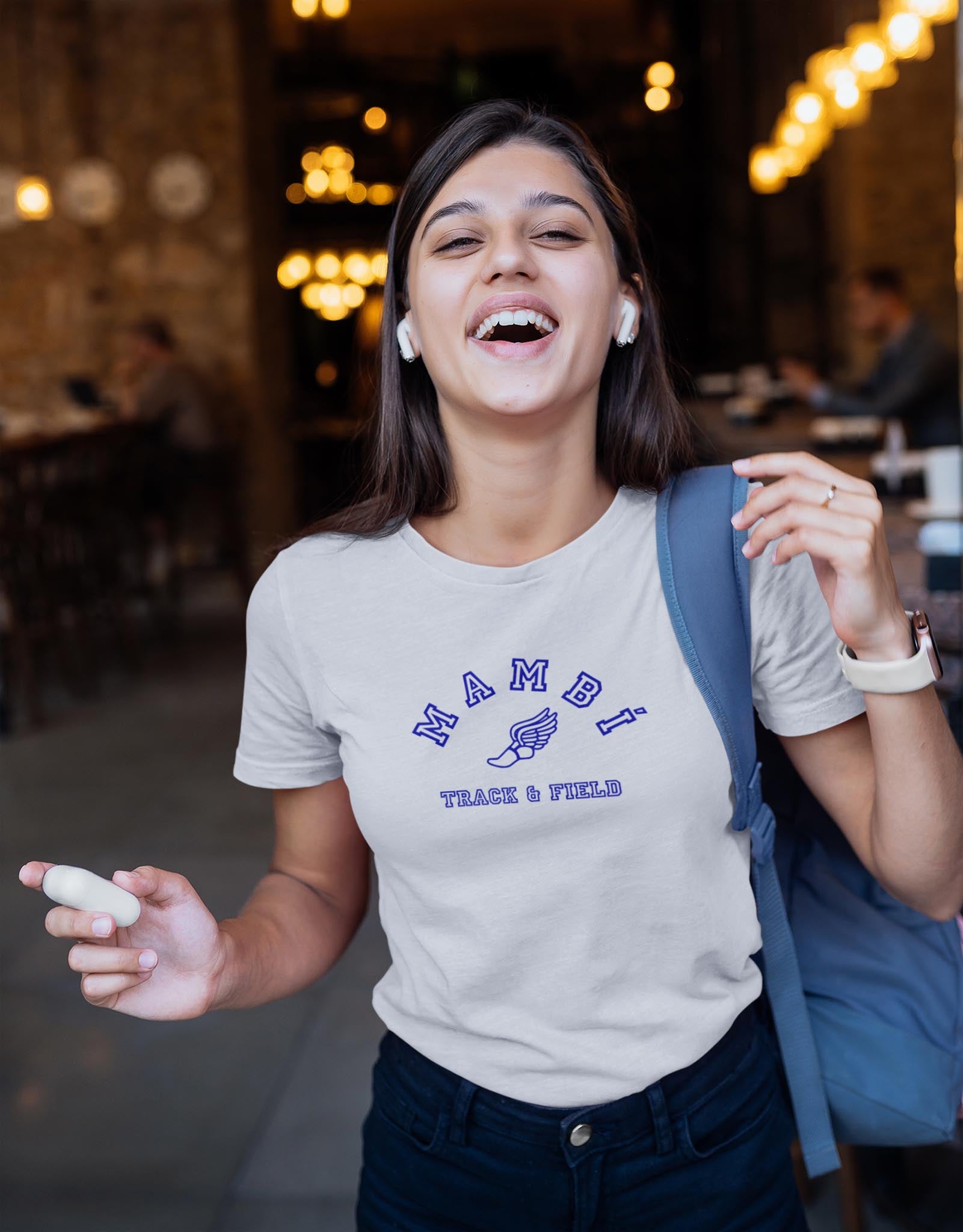 A young woman smiles while holding wireless earbuds and wearing a light gray Mambi Track & Field T-shirt, with a blue backpack over one shoulder, inside a warmly lit cafe or restaurant.