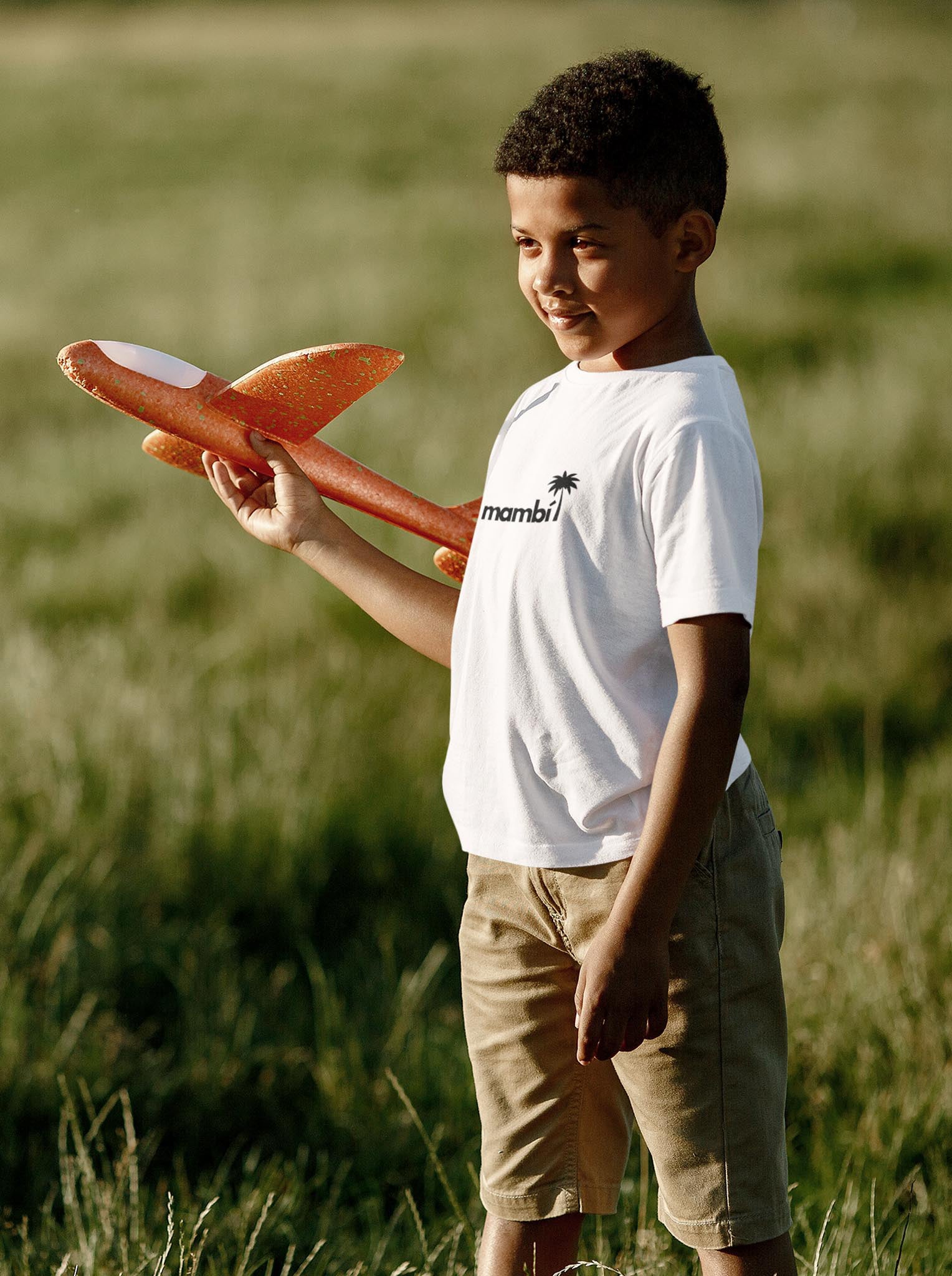 A young boy stands in a grassy field, holding an orange toy glider airplane. He wears a white t-shirt and khaki shorts, smiling softly as he looks at the plane, bathed in warm sunlight.