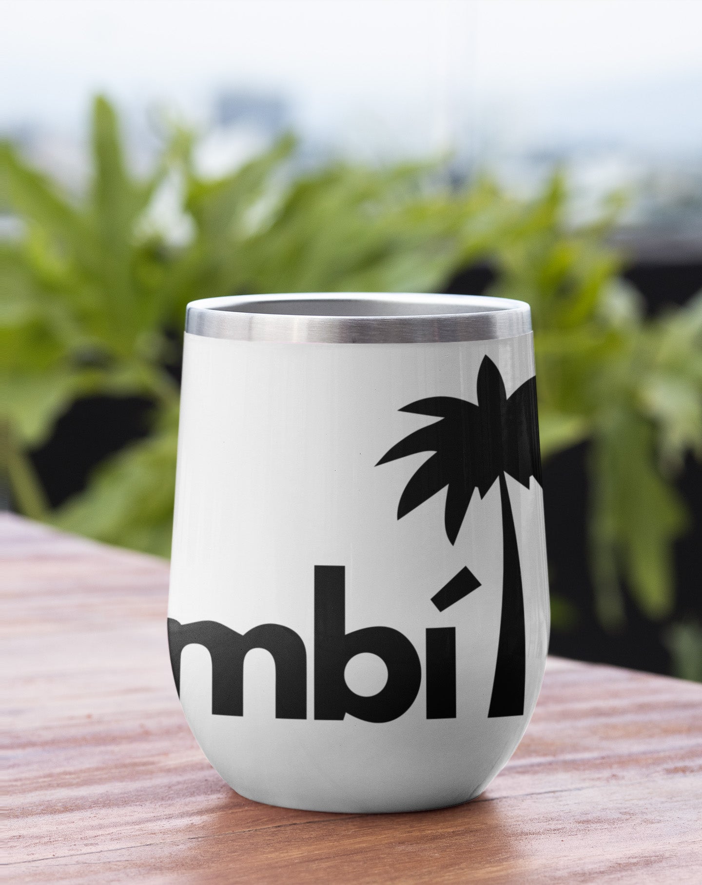 A white tumbler with a black palm tree graphic and partial bold text reading mbi sits on a wooden surface with green, leafy plants blurred in the background.