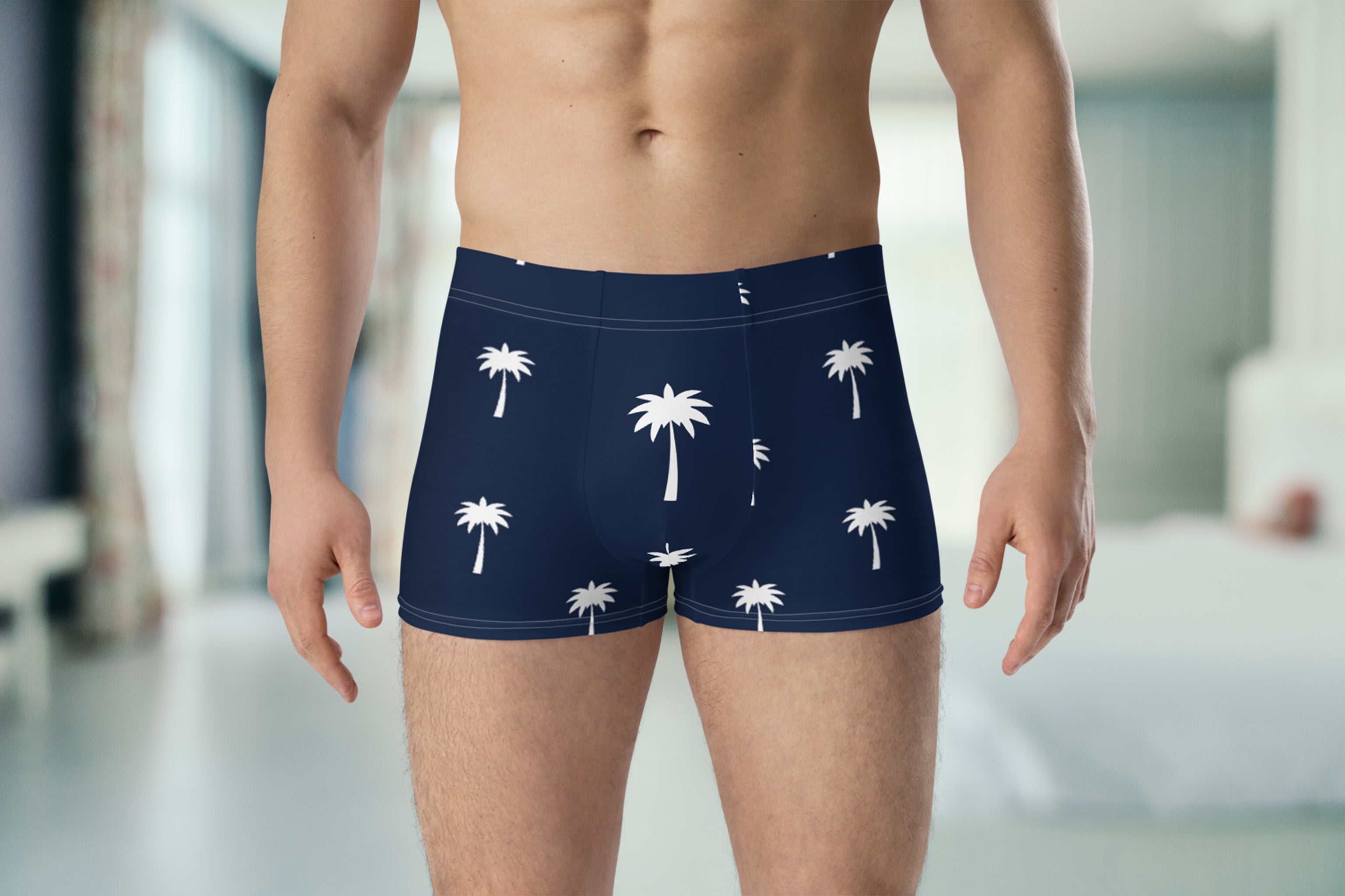 A man stands indoors wearing navy blue boxer briefs with a white palm tree pattern; his torso and upper legs are visible, with a blurred bedroom in the background.
