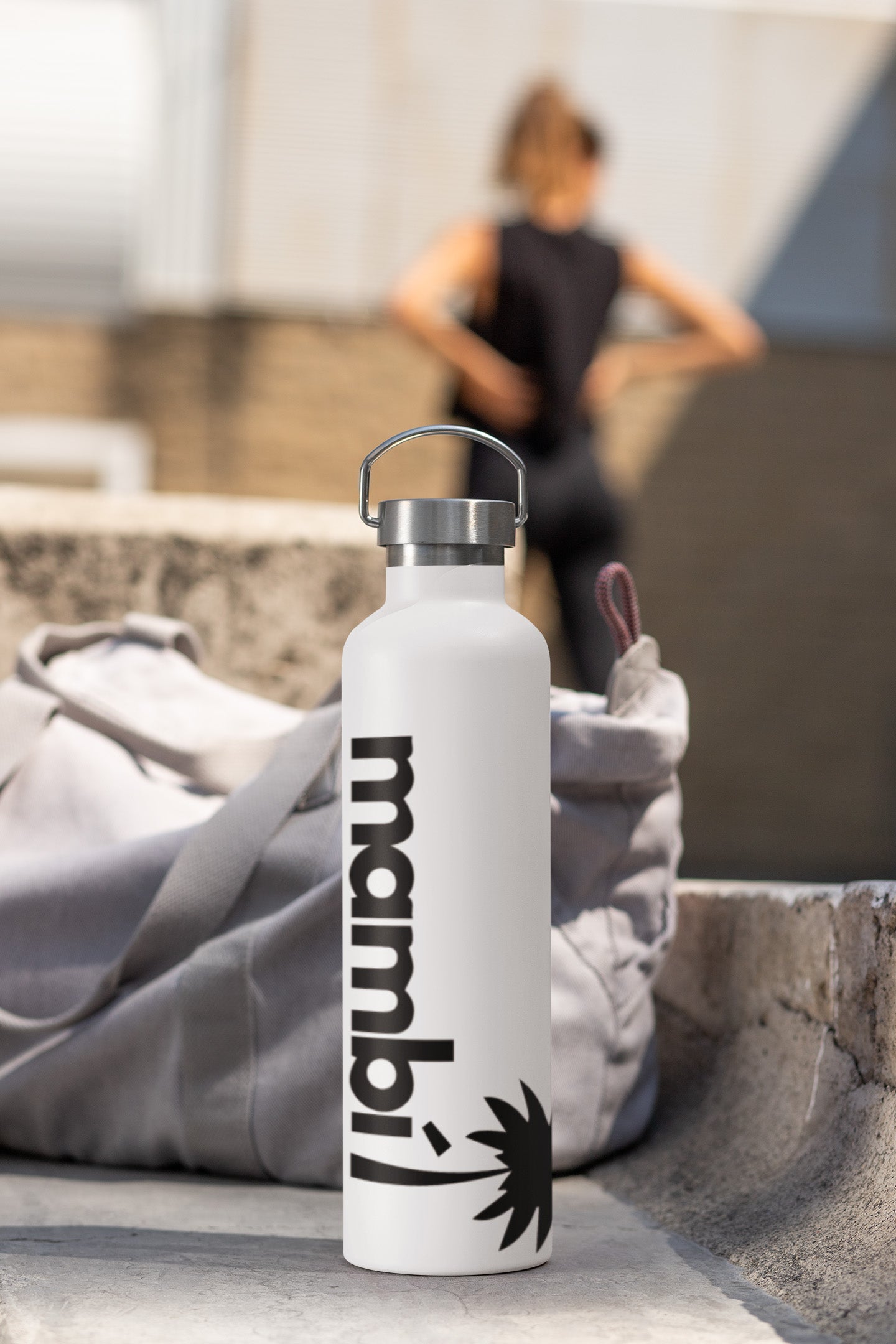 A white water bottle with a metal lid and mambí written on it rests on a light gray gym bag. In the blurred background, a person stands outside with hands on hips.