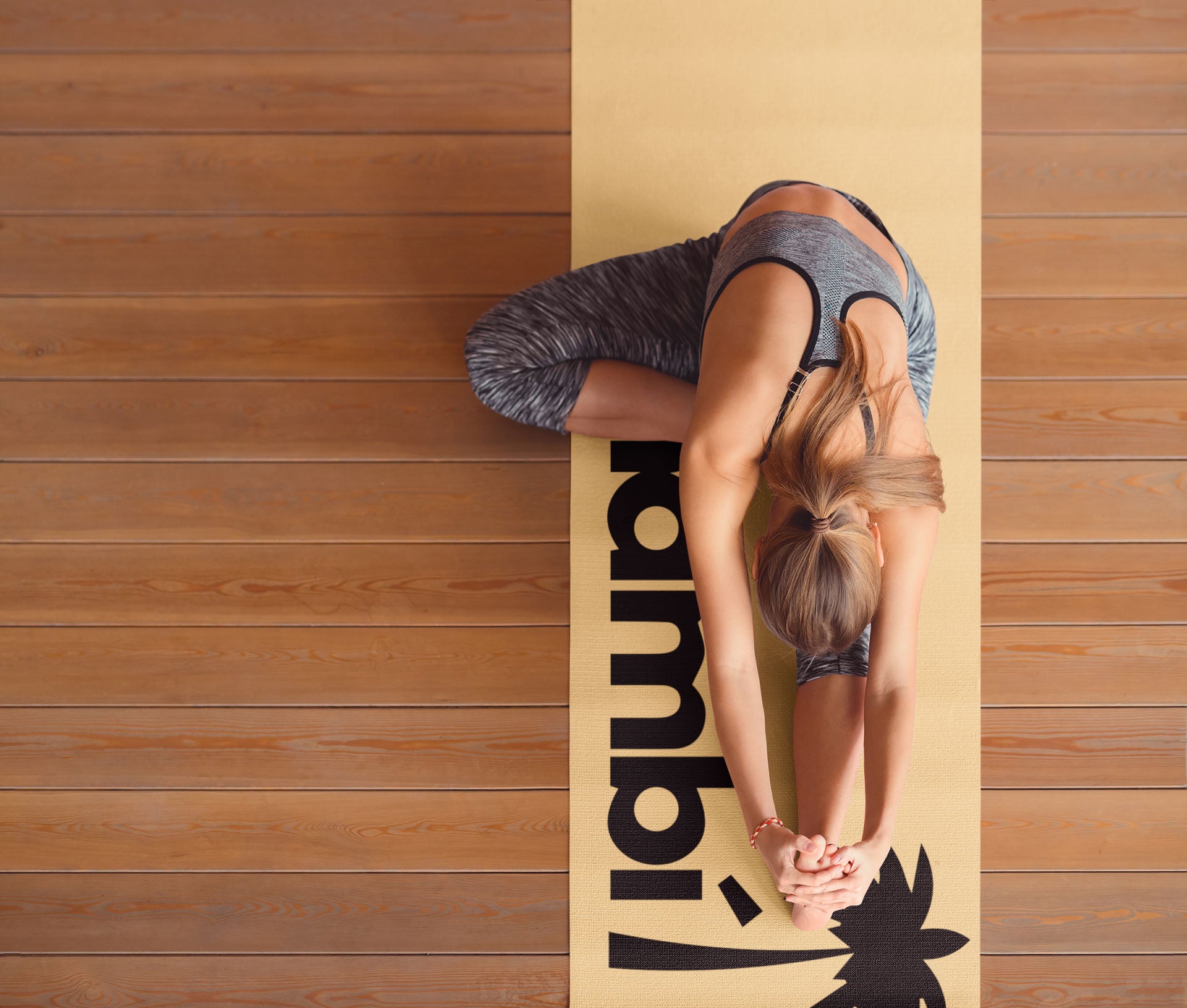 A woman wearing athletic clothes is sitting on a yellow yoga mat in a forward bend stretch, reaching her arms out in front of her, on a wooden floor.
