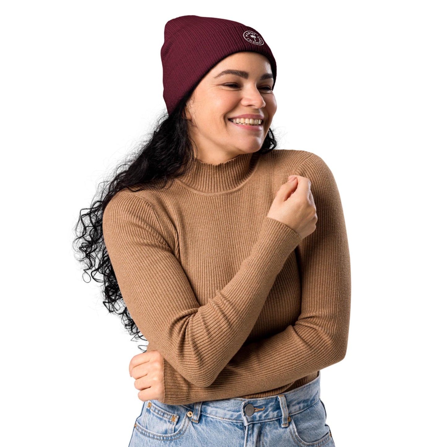 Woman with long curly dark hair wearing a maroon beanie, brown ribbed sweater, and light blue jeans, smiling with eyes closed and arms crossed, standing against a plain white background in a relaxed Mambí style.