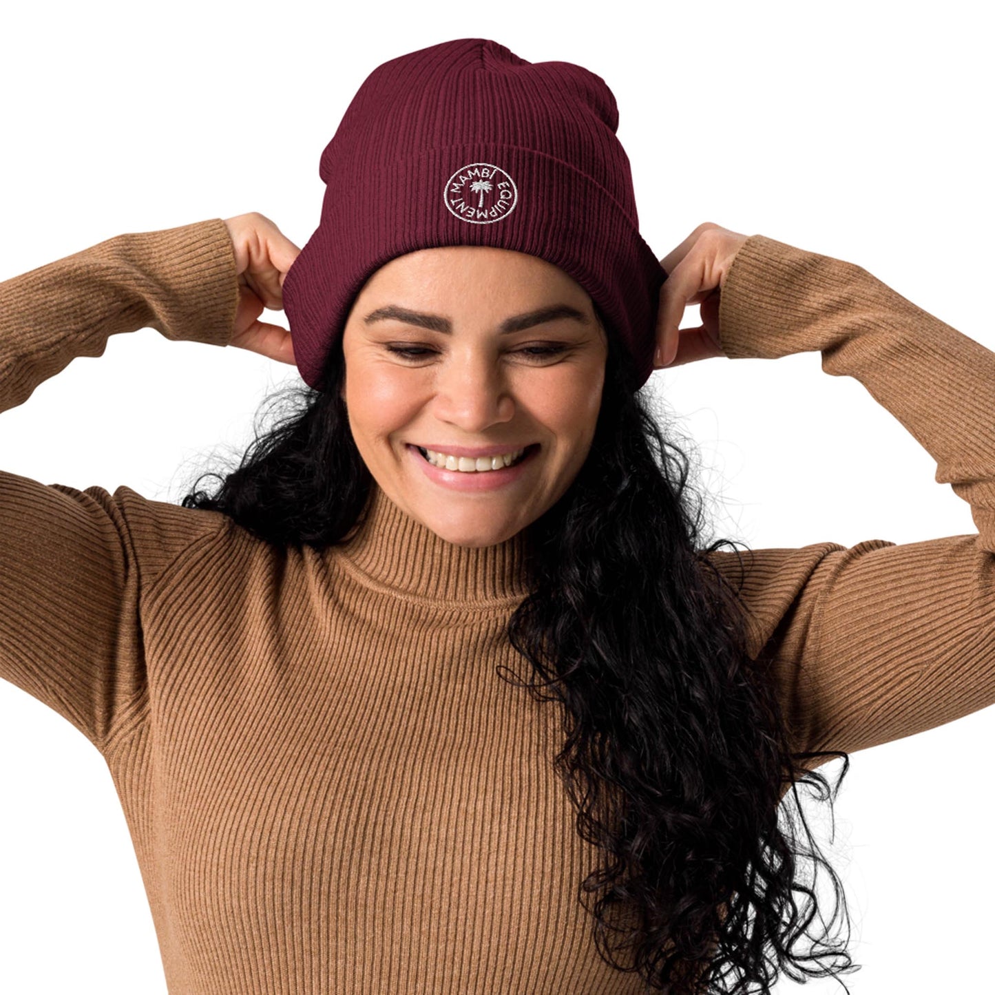 Smiling woman with long curly dark hair wears a ribbed brown sweater and a maroon Mambí beanie, adjusting the hat with both hands. The beanie features a small white circular logo on the front. The background is white.