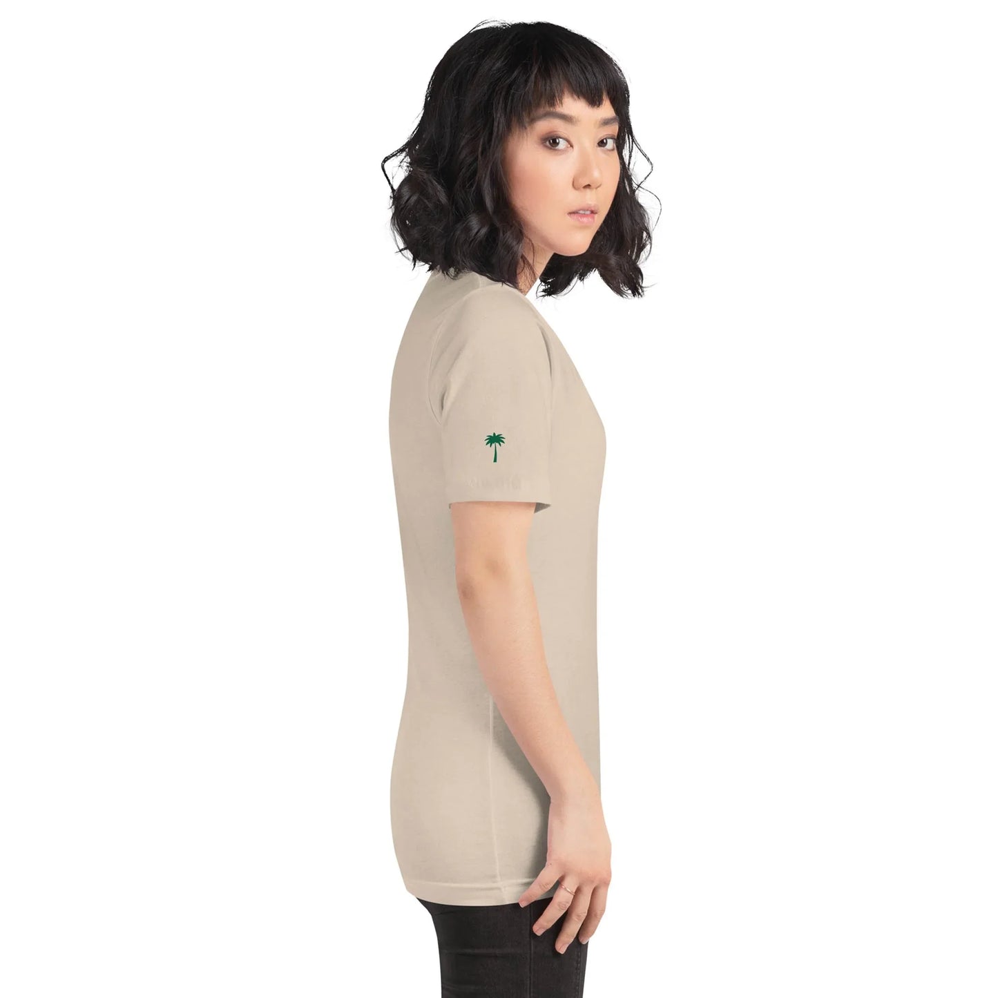 A woman with wavy black hair, wearing Printfuls unisex Cuban Bros graphic tee from the Neighborhood Collection and black pants, stands in profile facing left. The beige shirt has a small green palm tree on the sleeve against a plain white background.