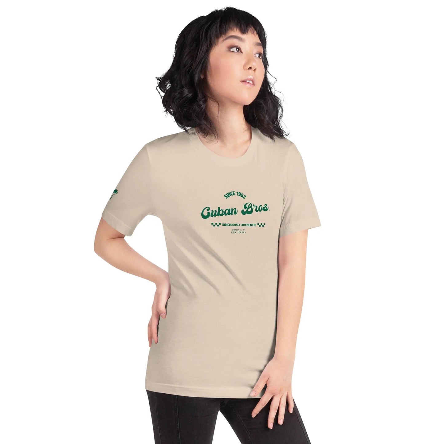 A woman with medium-length dark hair wears the Printful Cuban Bros Graphic Tee (Unisex) featuring green lettering, standing with one hand on her hip against a plain white background.