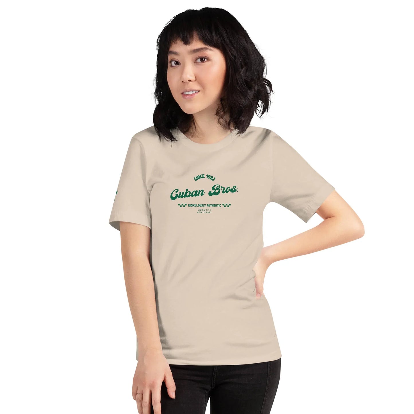A woman with shoulder-length dark hair wears the Printful Cuban Bros Graphic Tee | Unisex from the Neighborhood Collection, featuring Since 1998 Cuban Bros in green, standing with one hand on her hip and smiling at the camera.
