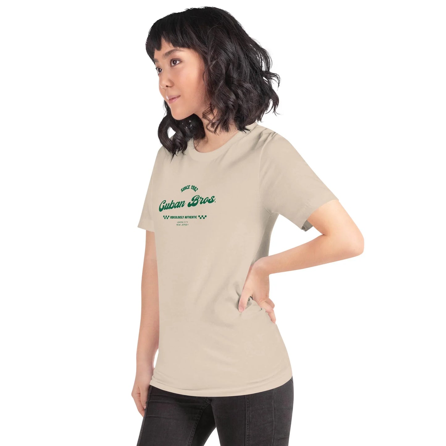 A woman with shoulder-length dark hair and bangs stands sideways in a light beige Cuban Bros Graphic Tee from Printfuls Neighborhood Collection, paired with black pants, against a plain white background.