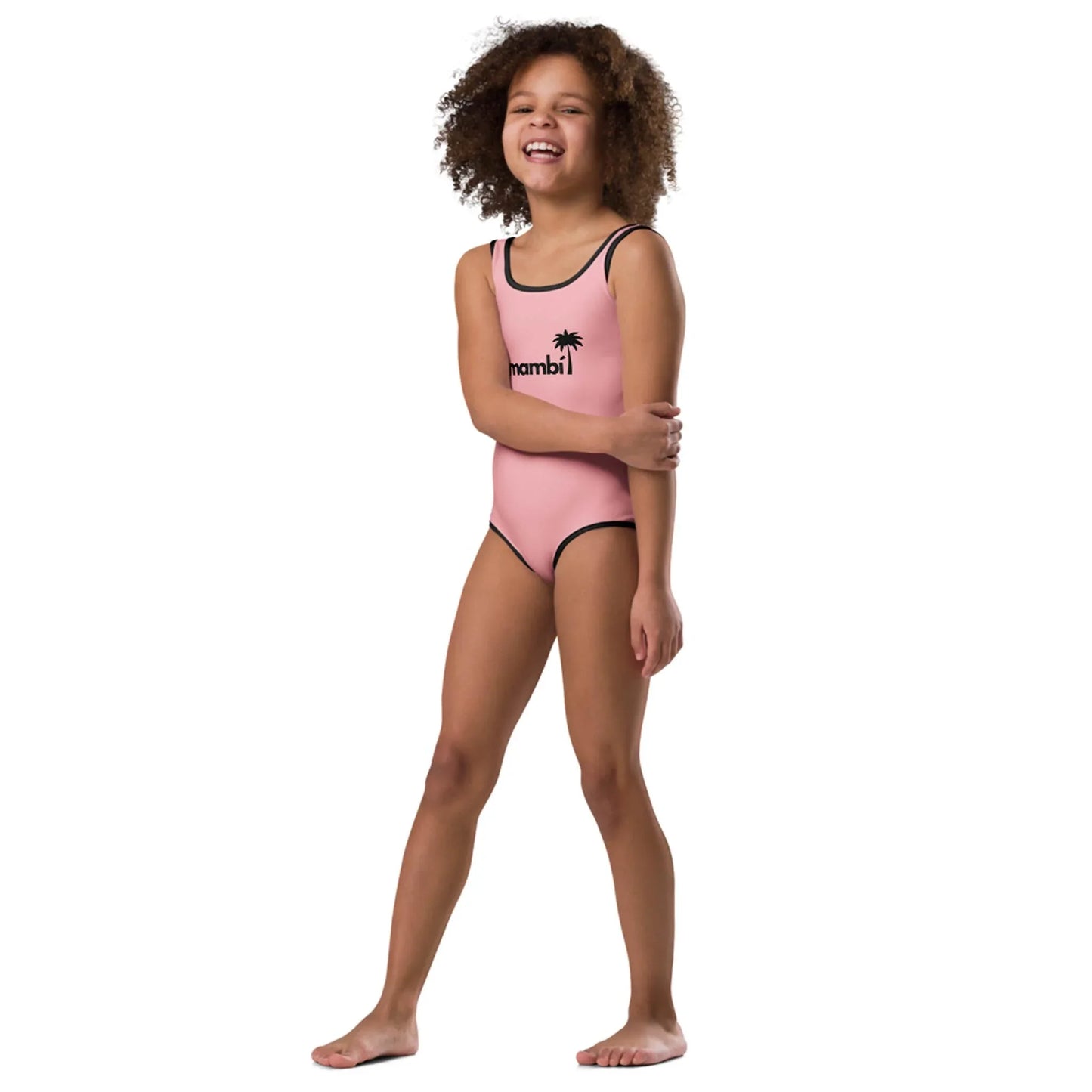 A smiling young girl with curly hair poses barefoot in Printful’s “Daisy” Classic One-Piece Swimsuit for toddlers and little kids, featuring vibrant prints, against a plain white background.