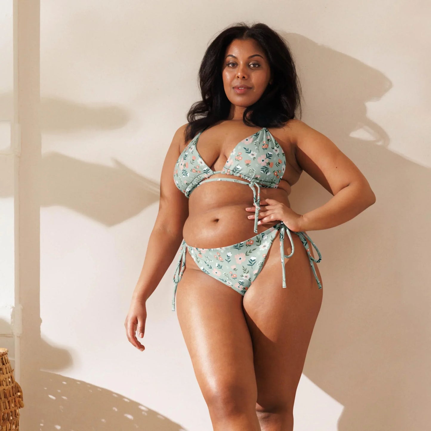 A woman with long dark hair poses indoors in natural sunlight, wearing the Daniella String Bikini Swimsuit by Printful, a light blue floral style made from recycled polyester.