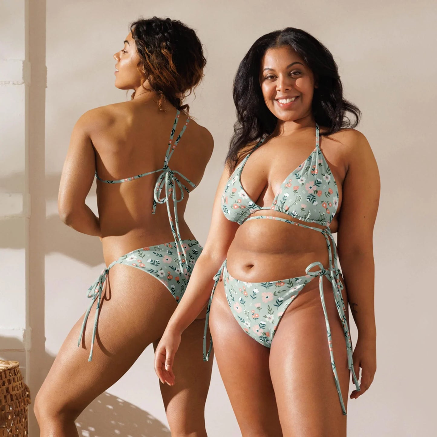 Two women model the Printful Daniella String Bikini Swimsuit in light blue floral—one facing forward, one showing the back. Made from recycled polyester with UPF 50+ protection, they stand in a brightly lit room with a neutral backdrop.