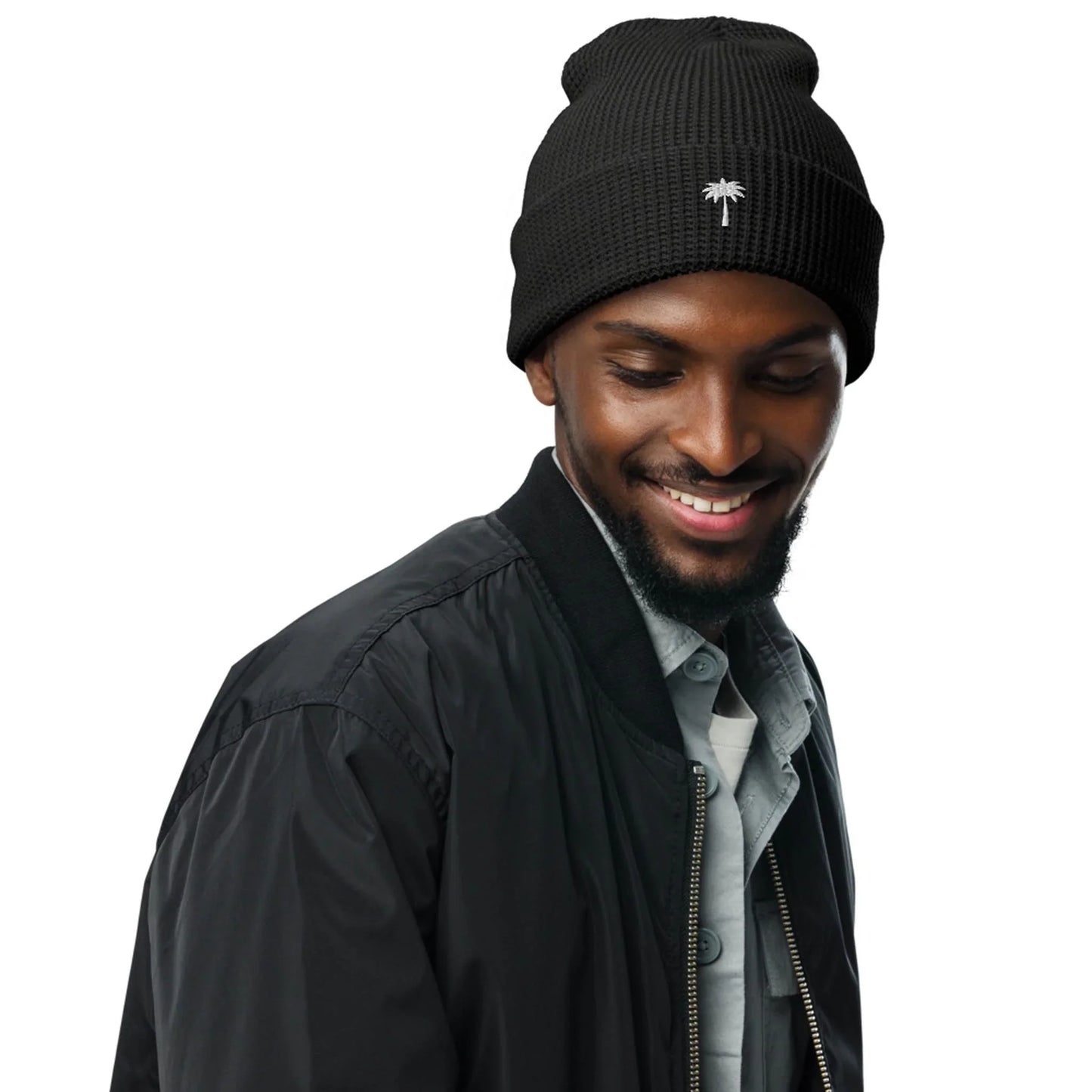 A man wearing a black beanie with a small palm tree logo, a black bomber jacket, and a light gray shirt, smiling and looking down to his left against a plain white background, gives off cool Mambí style.