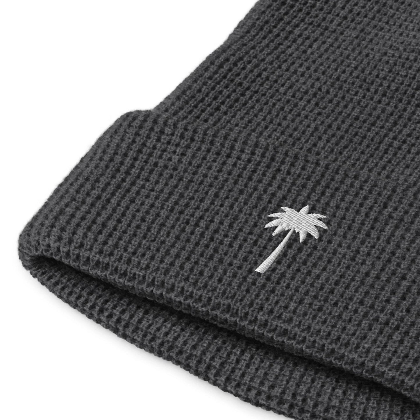 A close-up of a dark gray Mambí knitted beanie with a folded edge, featuring a small white embroidered palm tree on the cuff.