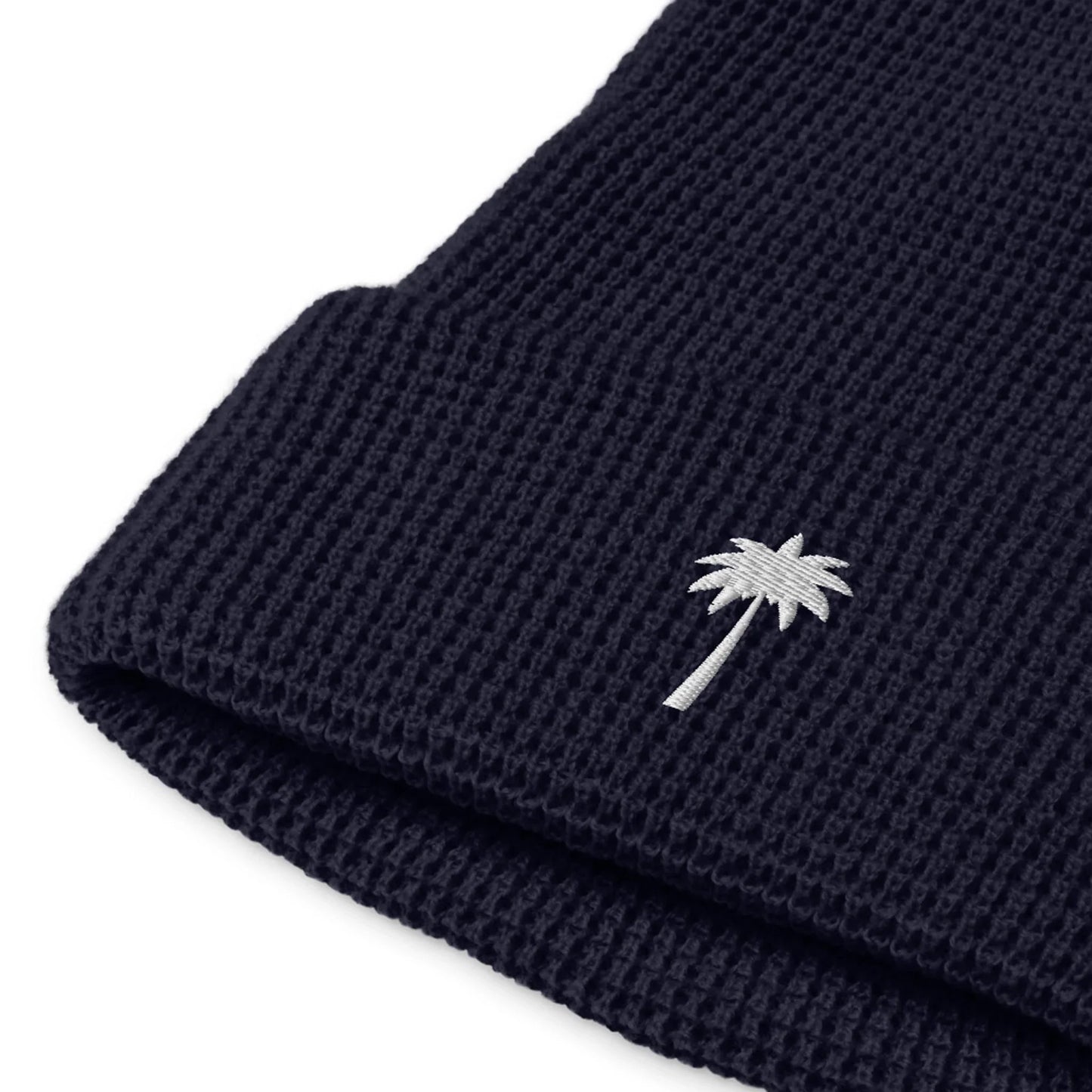 A close-up of a dark blue Mambí knit beanie featuring a white embroidered palm tree on the folded brim.