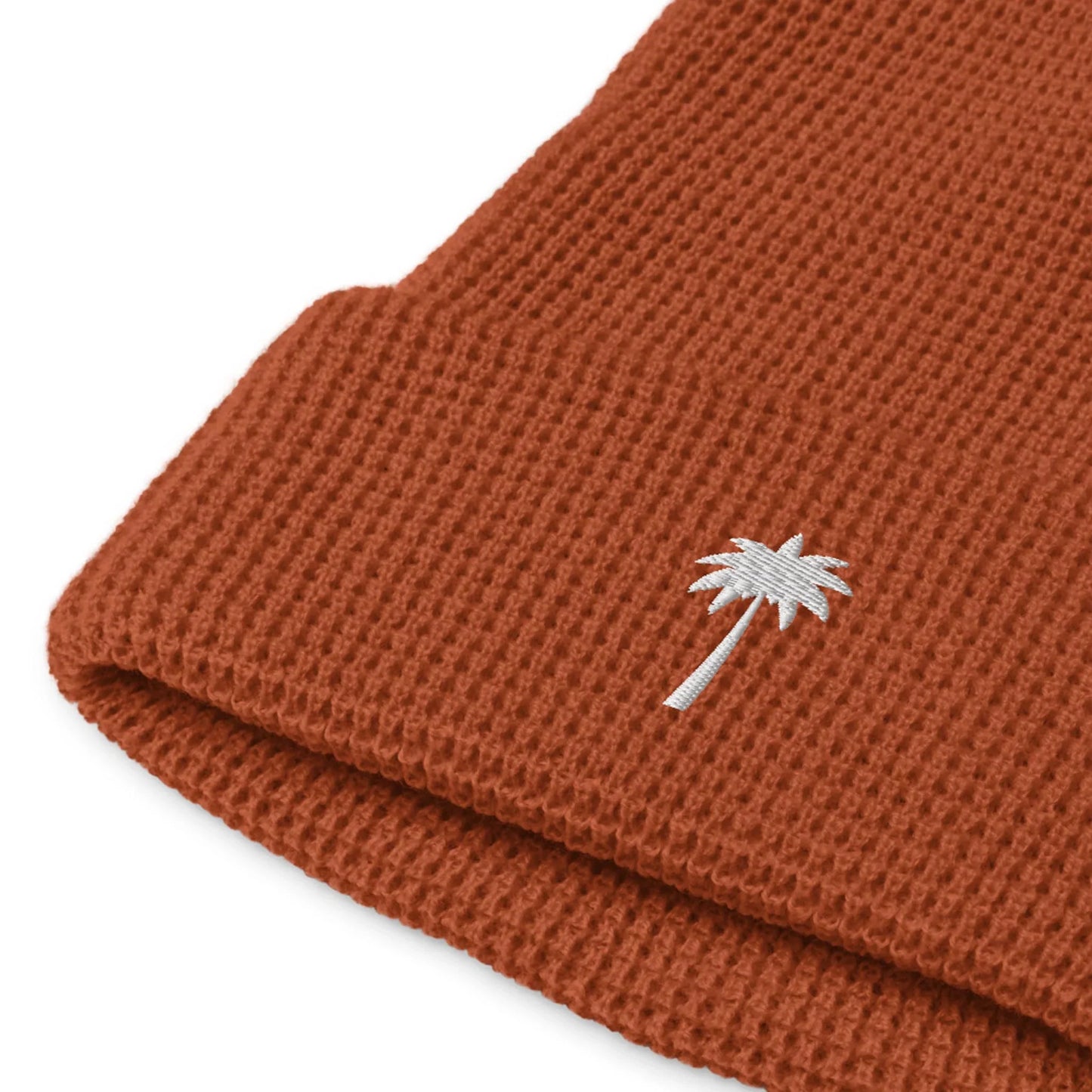Close-up of a rust-colored knit beanie with a folded brim, featuring a small white embroidered palm tree on the cuff—a stylish addition to the Mambí collection.