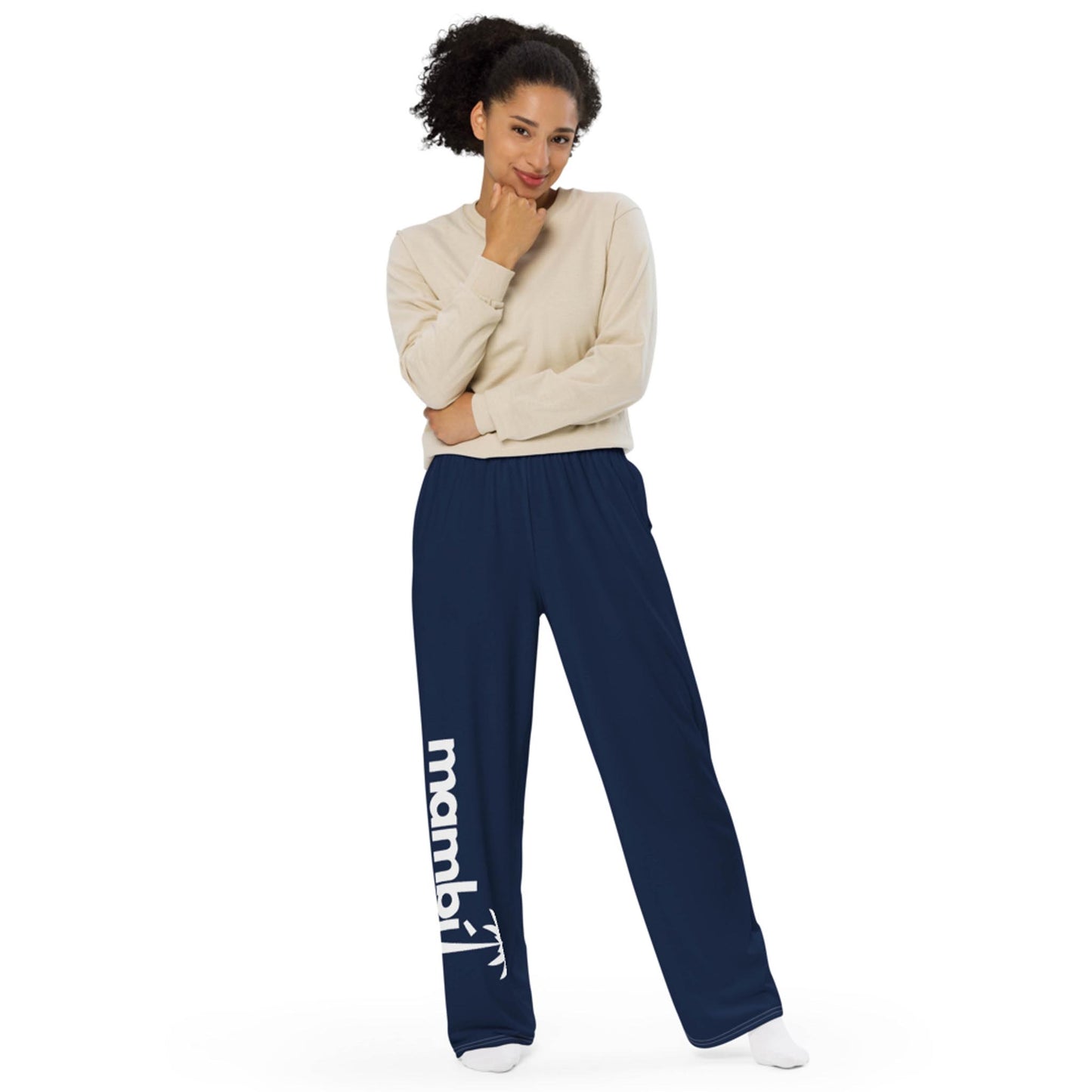 A woman stands smiling with one hand on her chin, wearing a beige long-sleeve shirt and loose navy blue Mambí pants featuring an arrow graphic in white on the left leg.