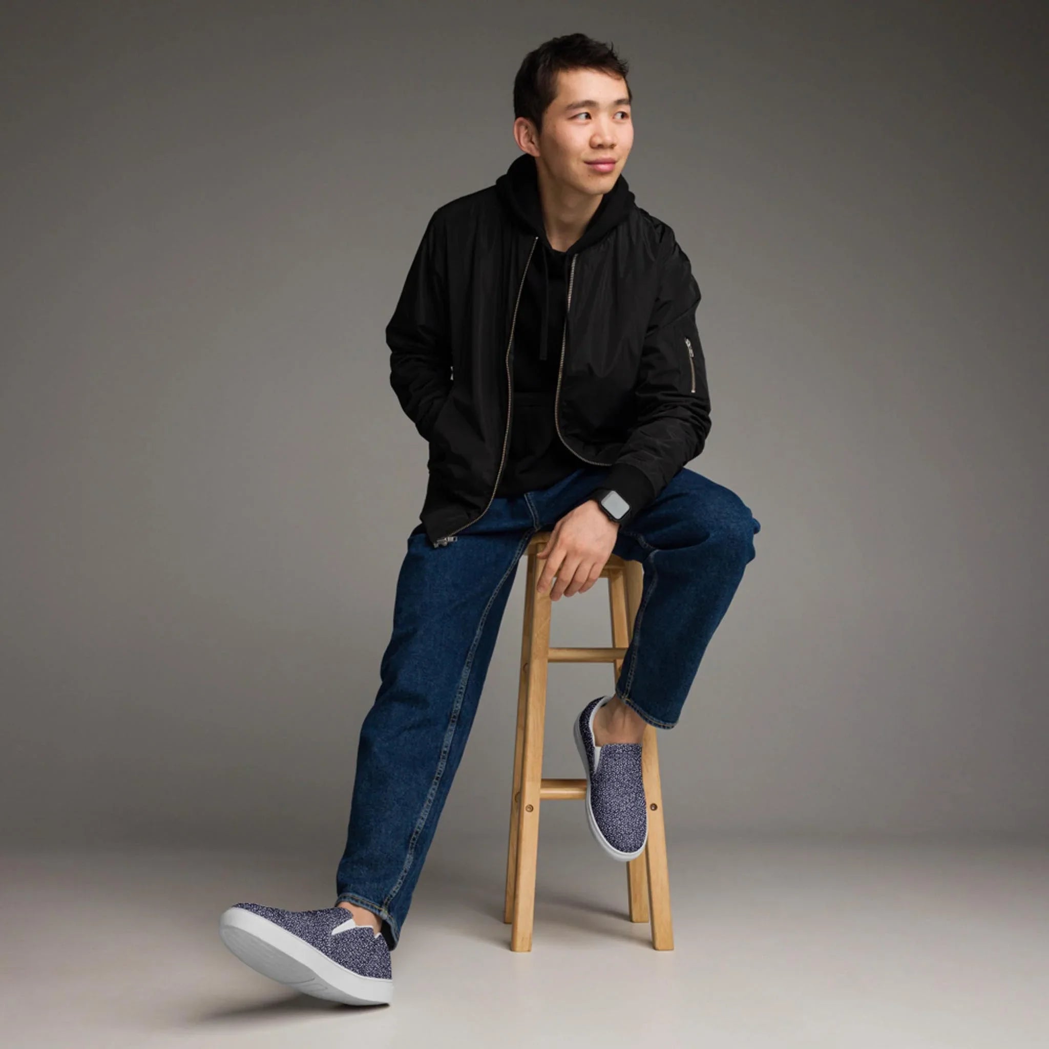 A young man in a black jacket and blue jeans sits casually on a wooden stool against a gray backdrop, wearing Printfuls Digno Handmade Slip-On Canvas Shoes for men and glancing to the side with a slight smile.