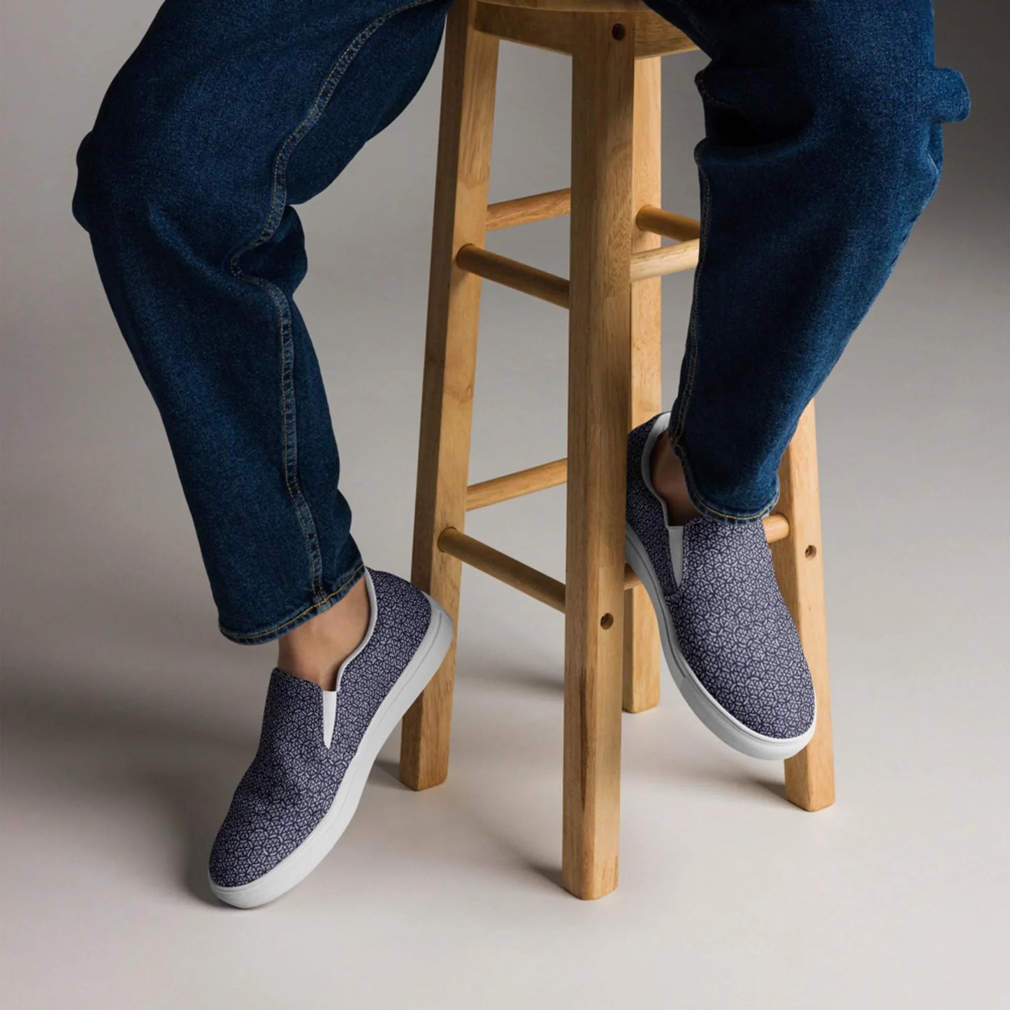 A person sits on a light wooden stool in blue jeans and Printfuls Digno Handmade Slip-On Canvas Shoes for men, with only the lower half visible, making a subtle fashion statement against a plain background.