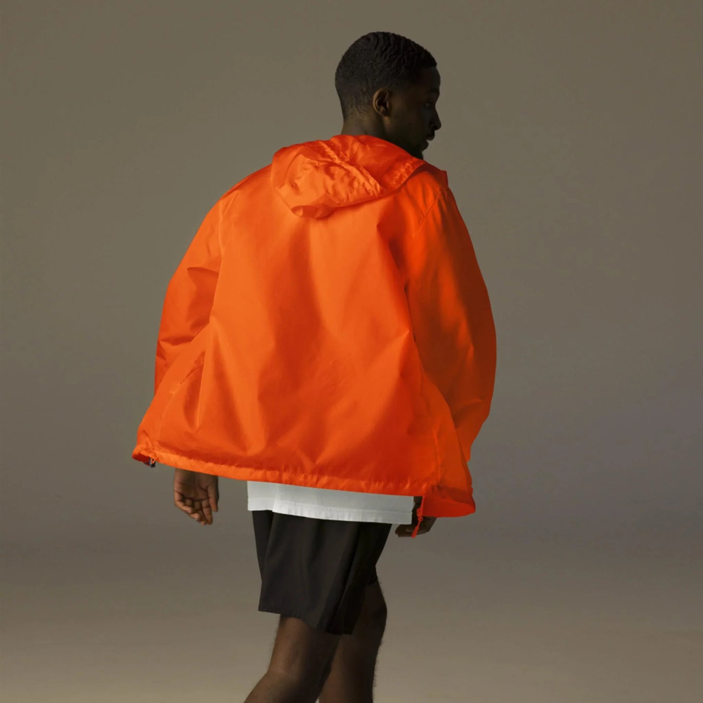 A person wearing a bright orange Mambí hooded jacket, white shirt, and black shorts is walking away from the camera against a neutral, softly lit background.