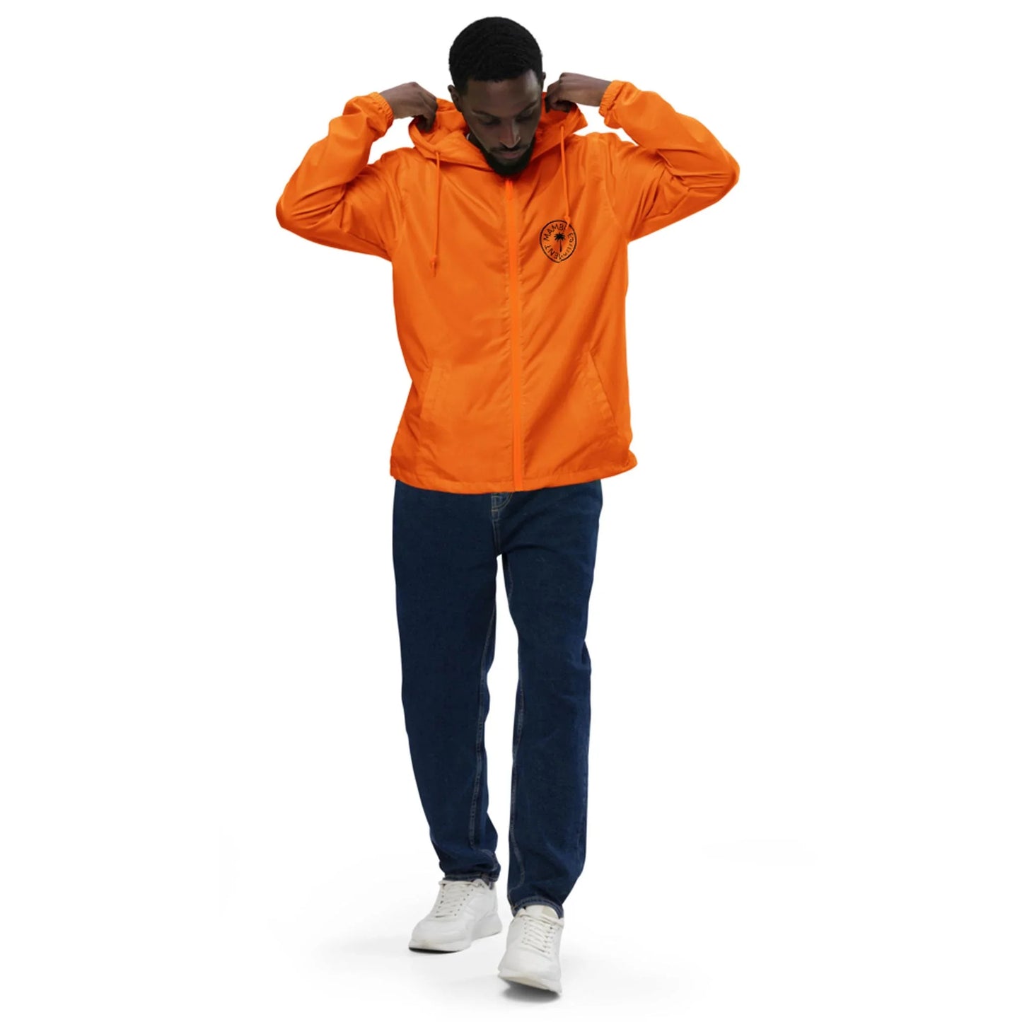 A man adjusts the hood of his bright orange Mambí jacket, featuring a small black emblem on the chest. He is wearing dark blue jeans and white sneakers, standing against a plain white background.