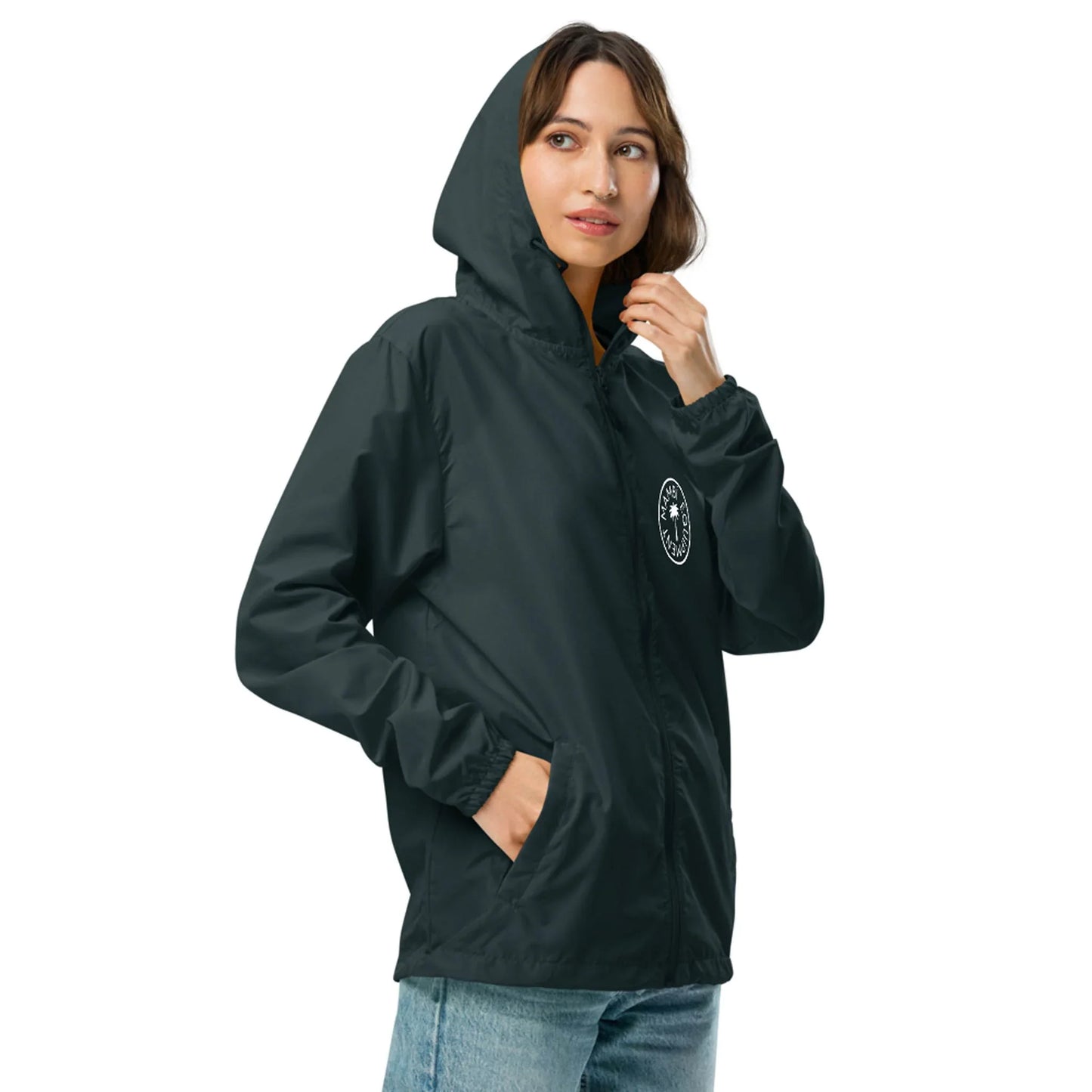 A woman wearing a dark green Mambí hooded jacket with a white circular logo on the chest stands with one hand near her face, looking slightly to the side. She has light brown hair and is wearing light blue jeans.