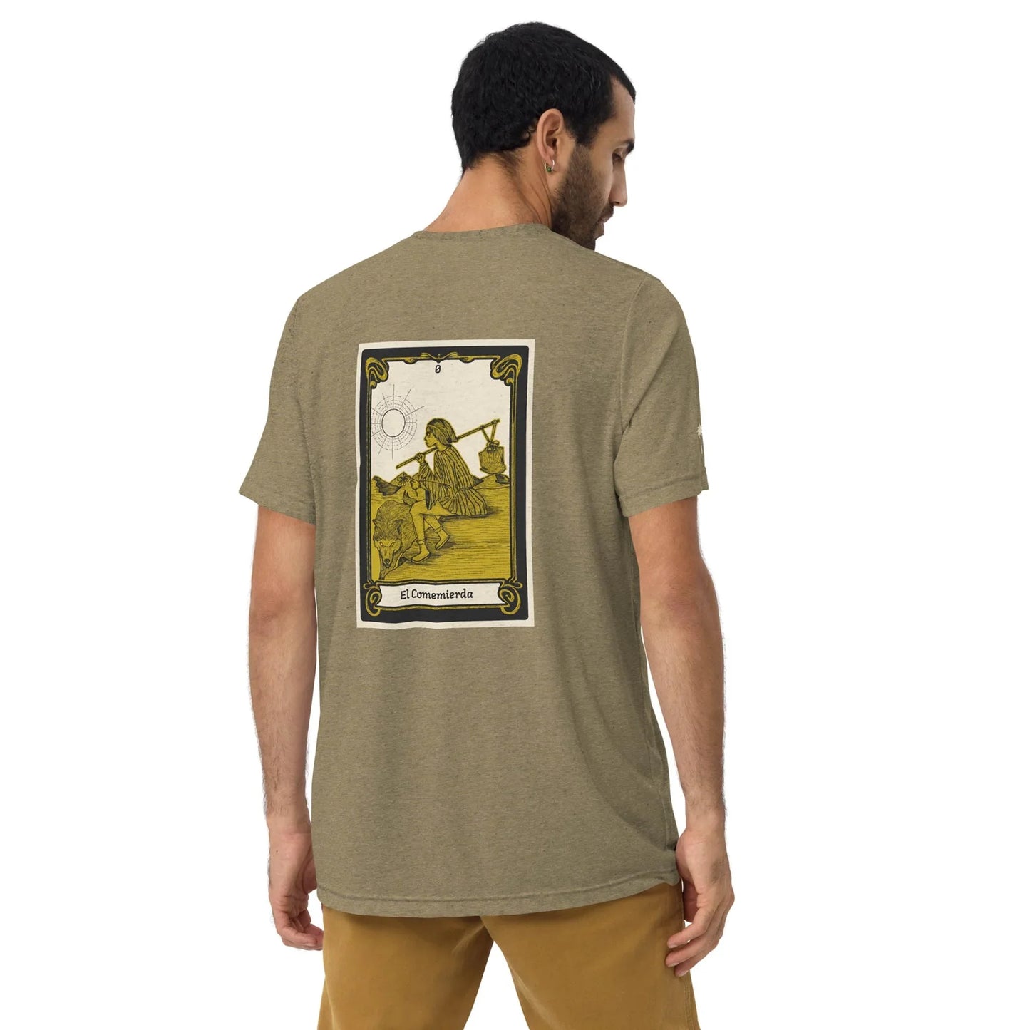 A man with short dark hair faces away, wearing the Printful El Comemierda Tri-Blend Graphic Tee, a comfy khaki unisex shirt with a bold card-style graphic of a person holding a spear and text El Comemierda on the back.