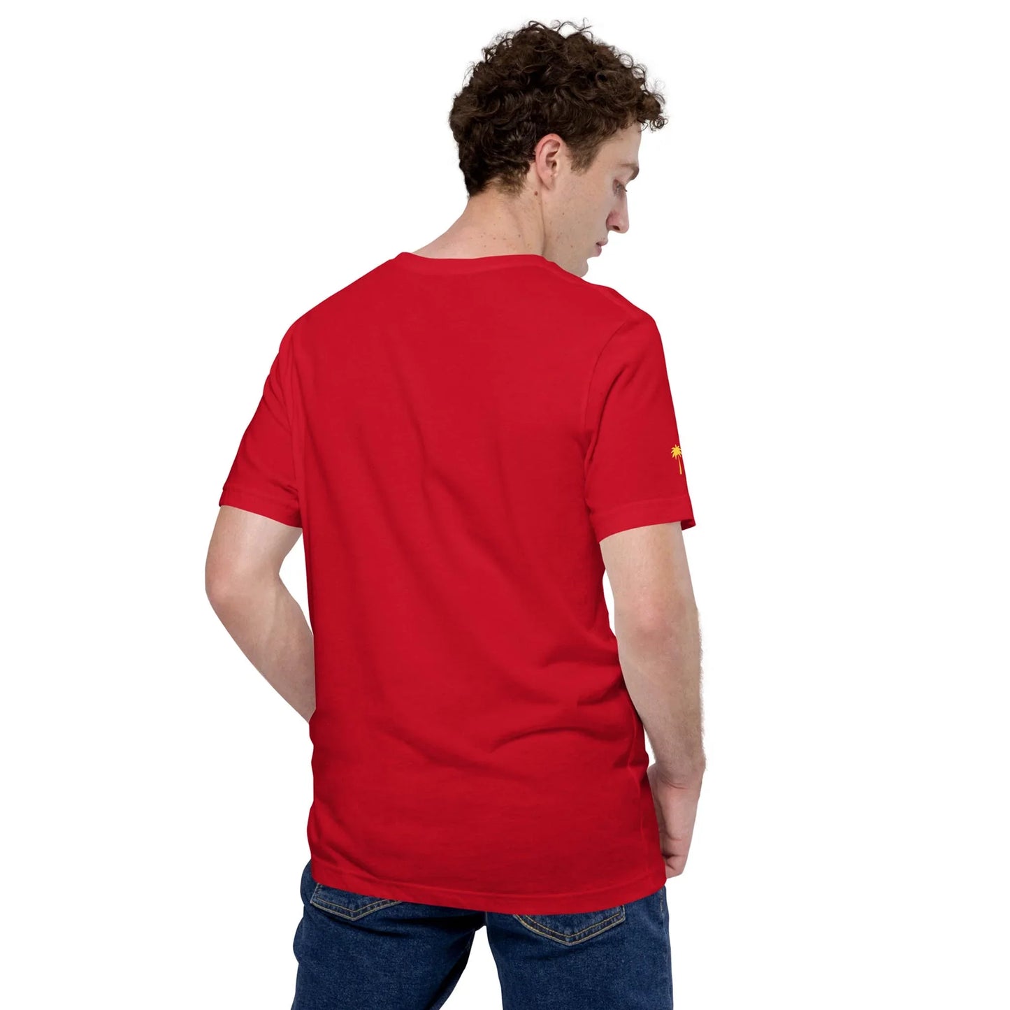 A man with curly brown hair, wearing a plain red Mambí T-shirt and blue jeans, stands with his back to the camera and looks over his left shoulder.