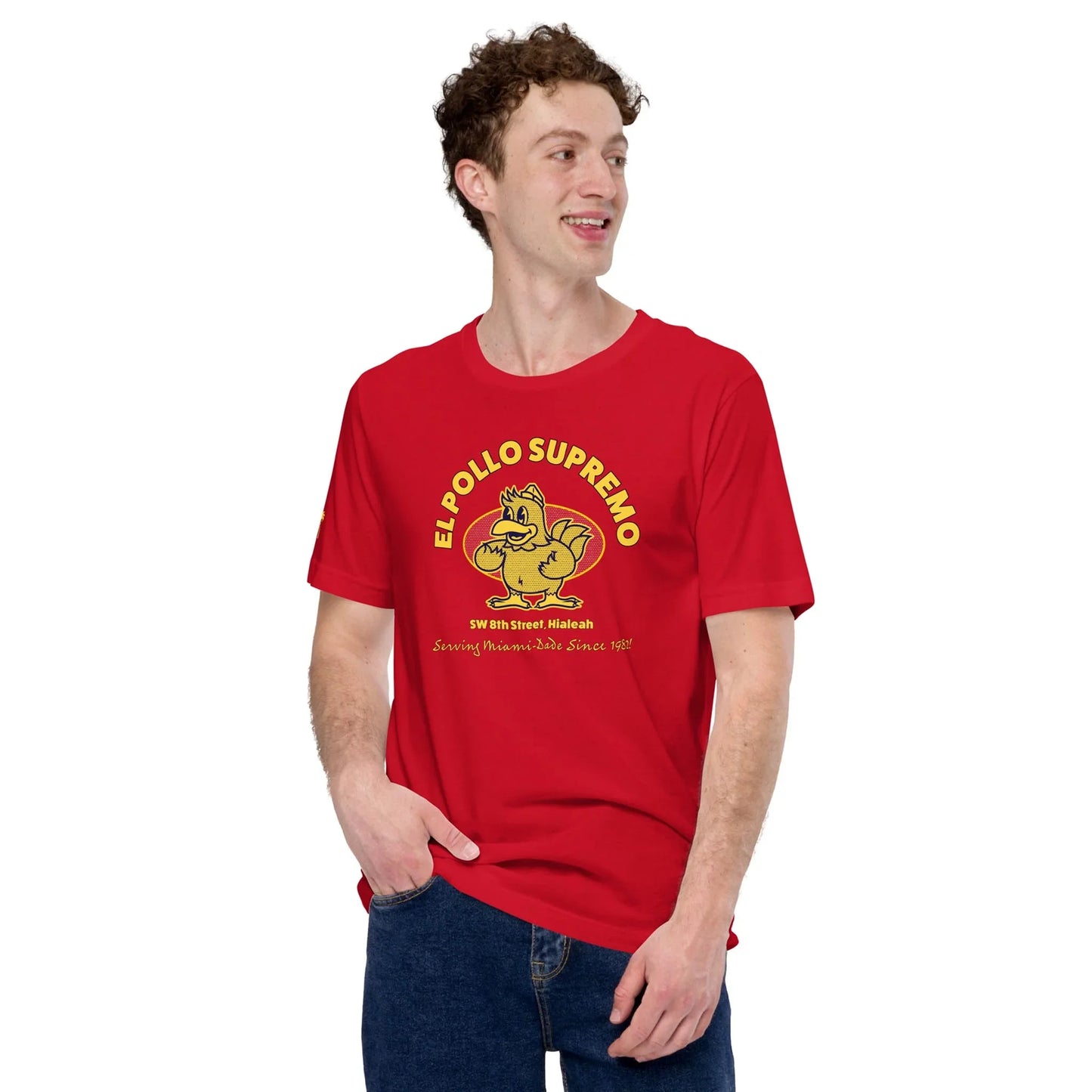 Young man with curly hair wearing a red El Pollo Supremo t-shirt featuring a cartoon chicken logo, standing with one hand in his pocket and looking to the side, smiling—a casual Mambí spirit shines through.