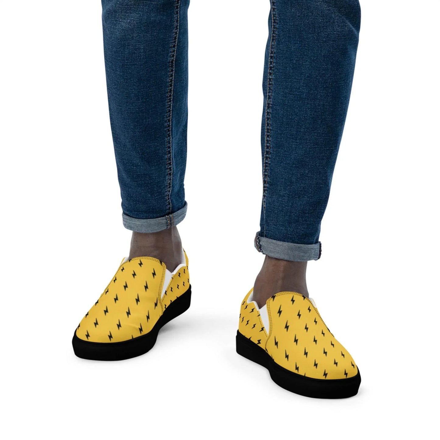 A person wearing rolled-up blue jeans and Printfuls Electra Handmade Slip-On Canvas Shoes for men, featuring a bold black lightning bolt pattern, stands out stylishly against a white background.