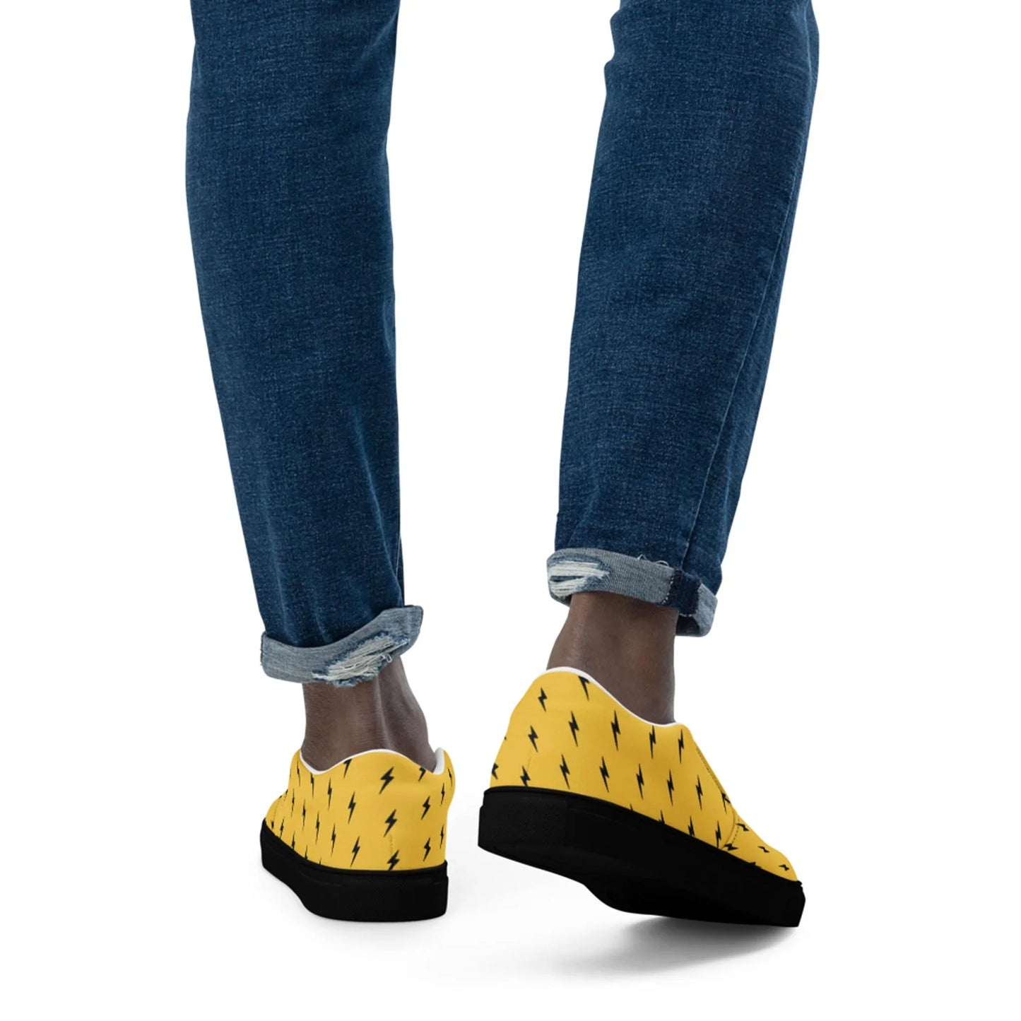 A person is seen from behind, wearing rolled-up blue jeans and Printfuls Electra Handmade Slip-On Canvas Shoes for men, featuring a bold black lightning bolt design—an eye-catching fashion statement against a white background.