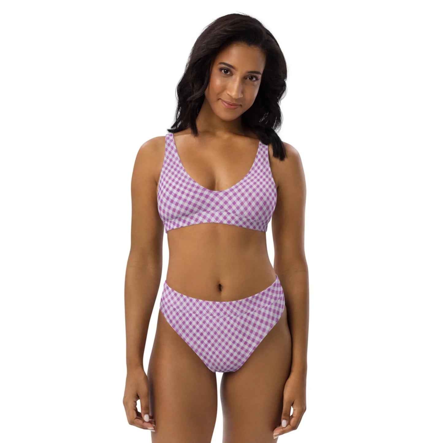 A woman with long dark hair wears a pink and white checkered Mambí bikini, standing and smiling softly against a plain white background.