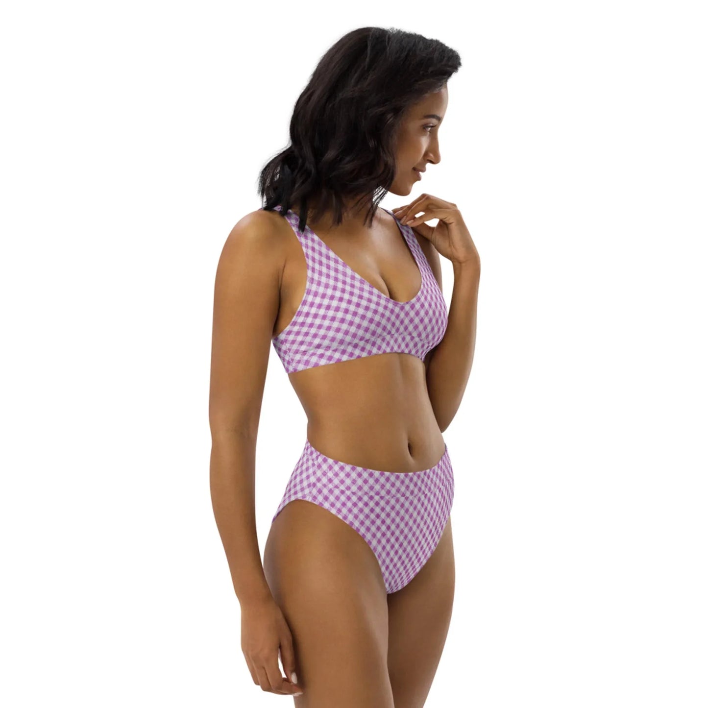 A woman in a purple and white checkered Mambí bikini stands against a plain white background, looking to her left with one hand raised near her chin.