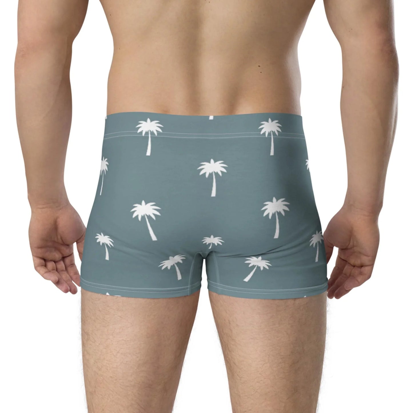 A man, shown from the back, wears Printfuls Emerson Boxer Brief for men in light blue with a white palm tree pattern. The boxer brief features a no-back-seam design and seamless fit for comfort, set against a plain neutral background.