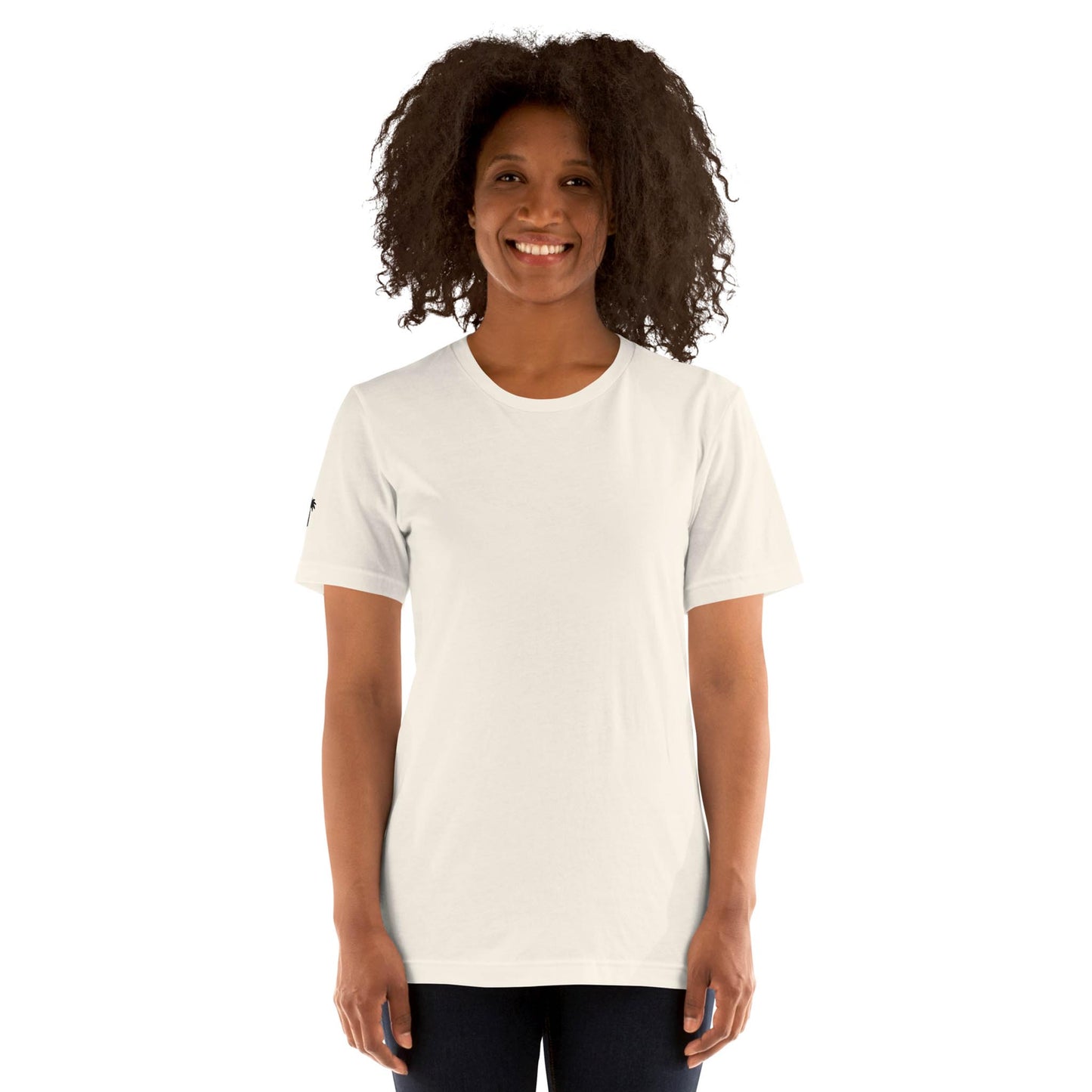 A woman with curly hair smiles while standing and wearing a plain white Mambí T-shirt and dark pants against a white background.