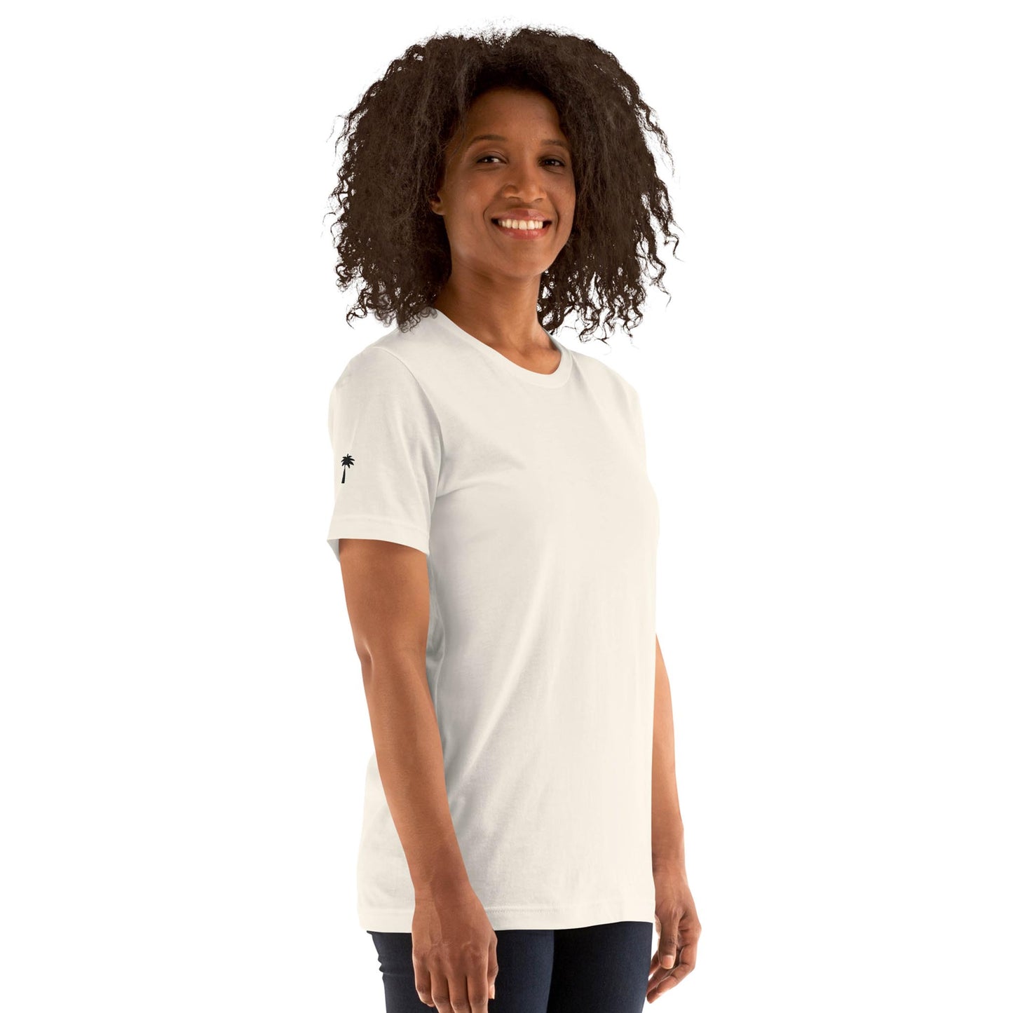 A woman with curly hair, wearing a plain white Mambí t-shirt and dark pants, smiles while standing at an angle against a white background.