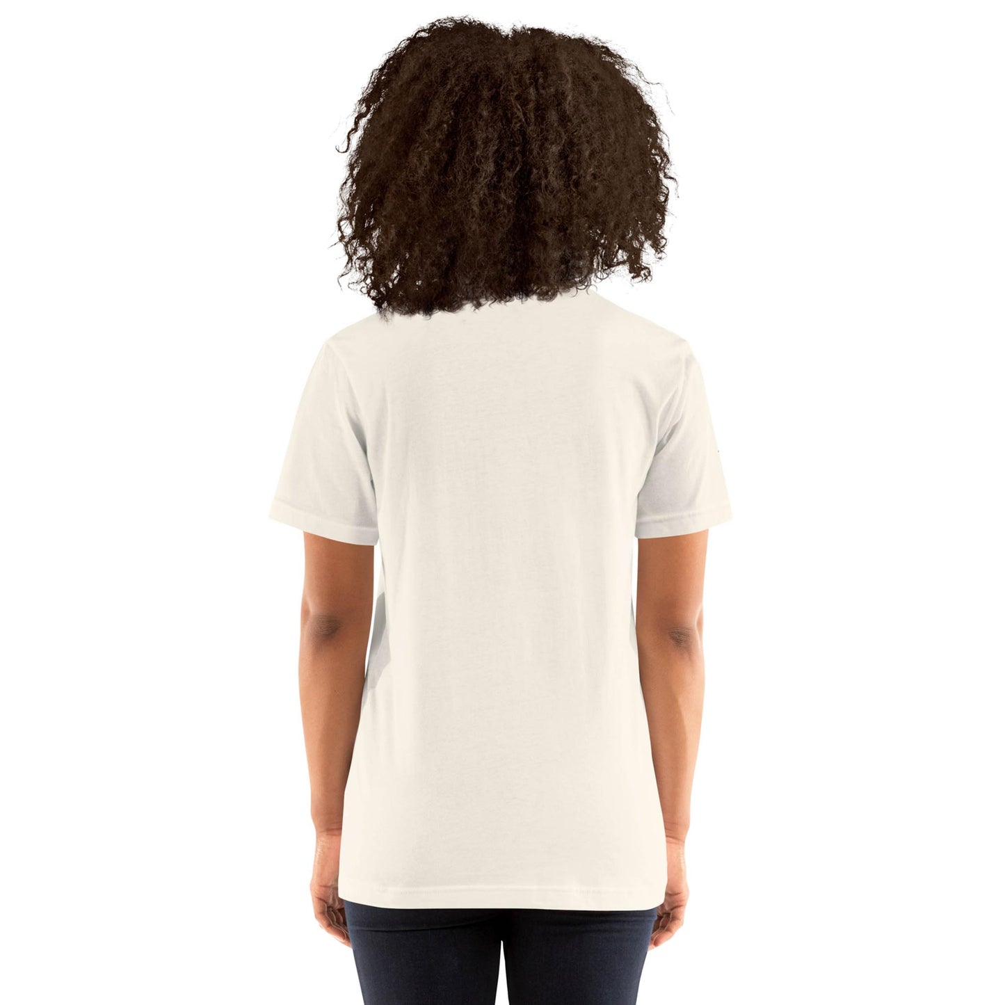 A person with curly hair is standing with their back to the camera, wearing a plain white Mambí short-sleeve t-shirt and dark jeans against a white background.
