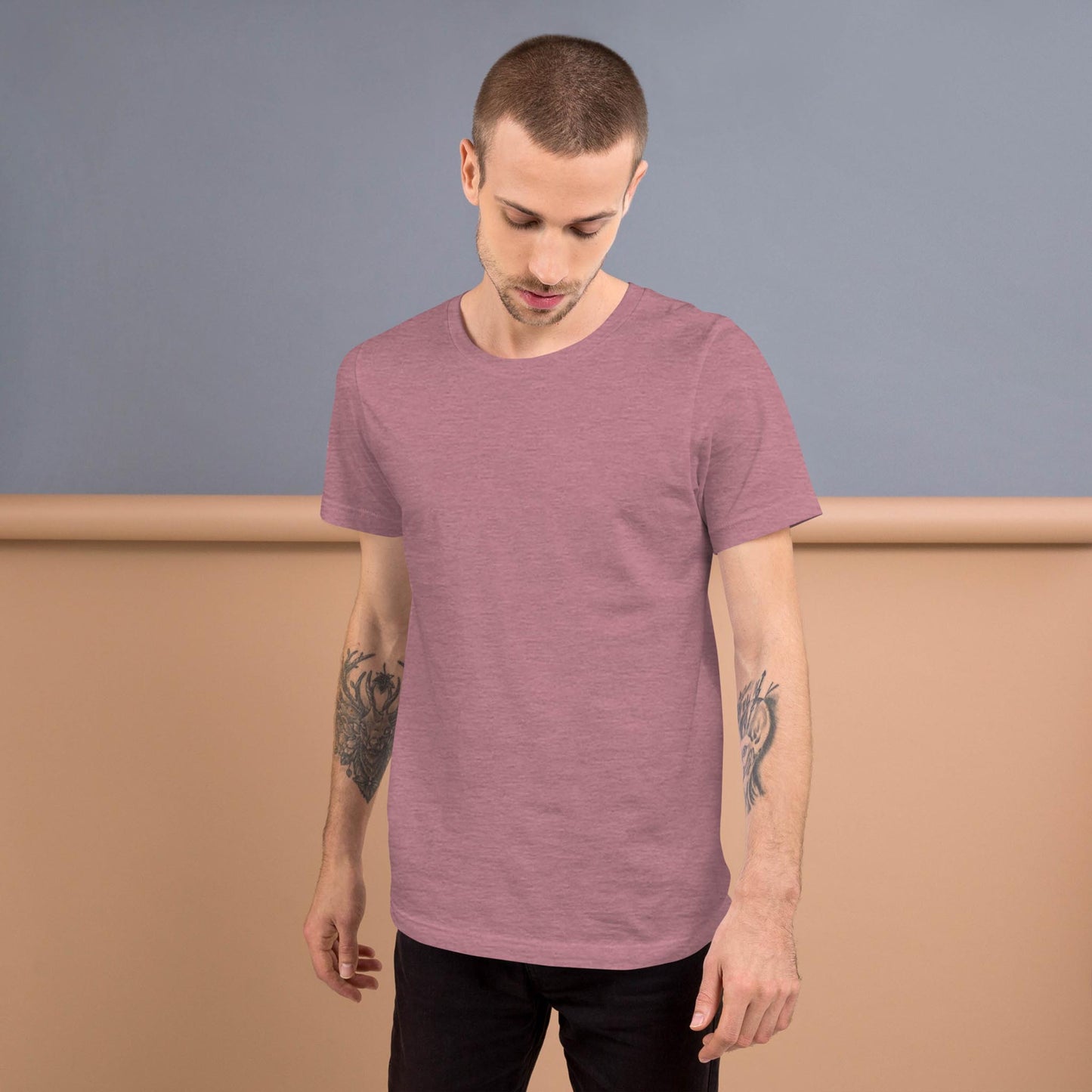 A man with short hair and arm tattoos is wearing a muted mauve Mambí t-shirt and black pants, standing against a two-tone beige and gray background, looking down with a neutral expression.