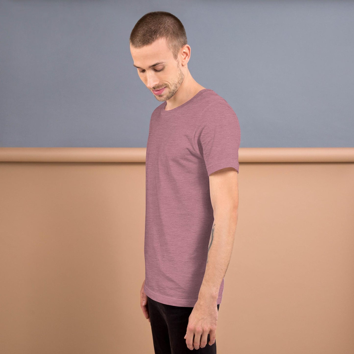 A man with short hair in a mauve Mambí t-shirt stands sideways, looking down, against a two-tone beige and gray background.