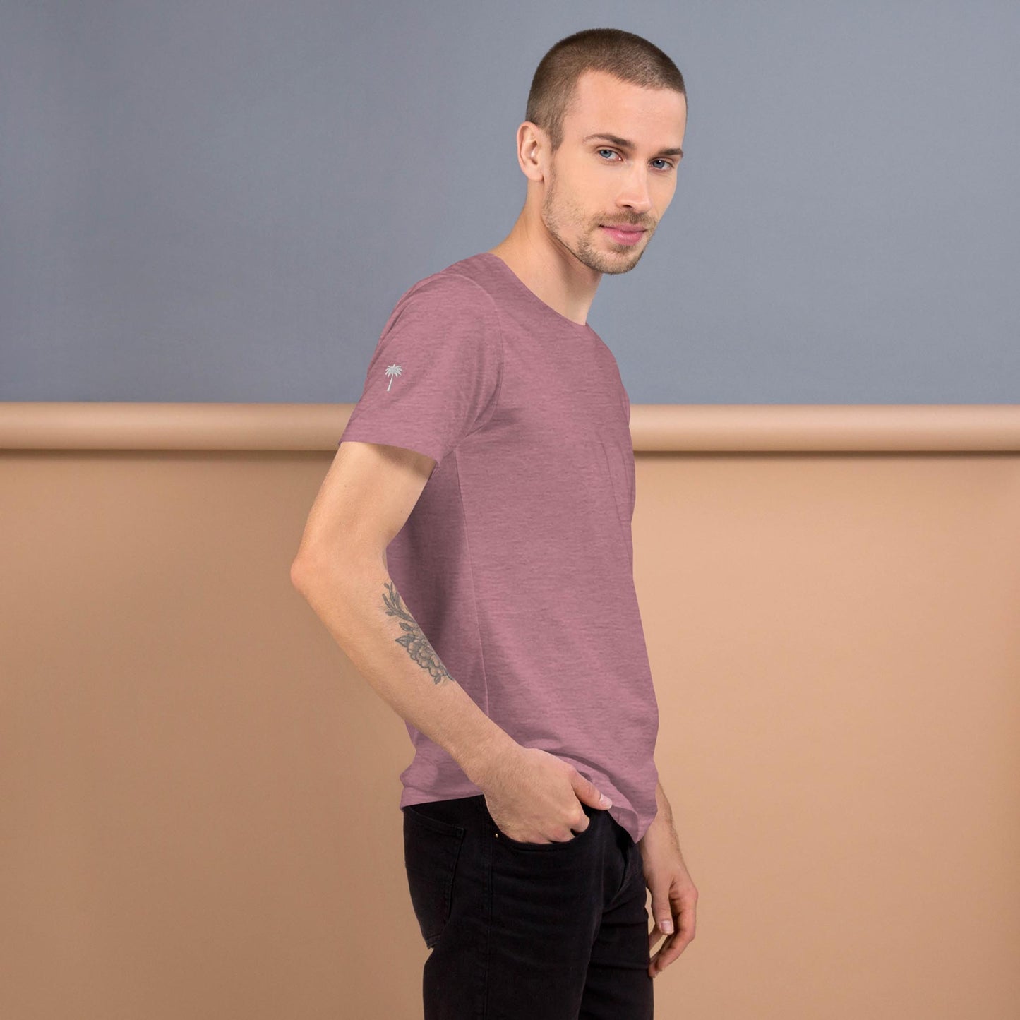 A man with short hair and a tattoo on his left forearm stands sideways, wearing a mauve Mambí t-shirt and black pants, against a two-tone beige and gray background.