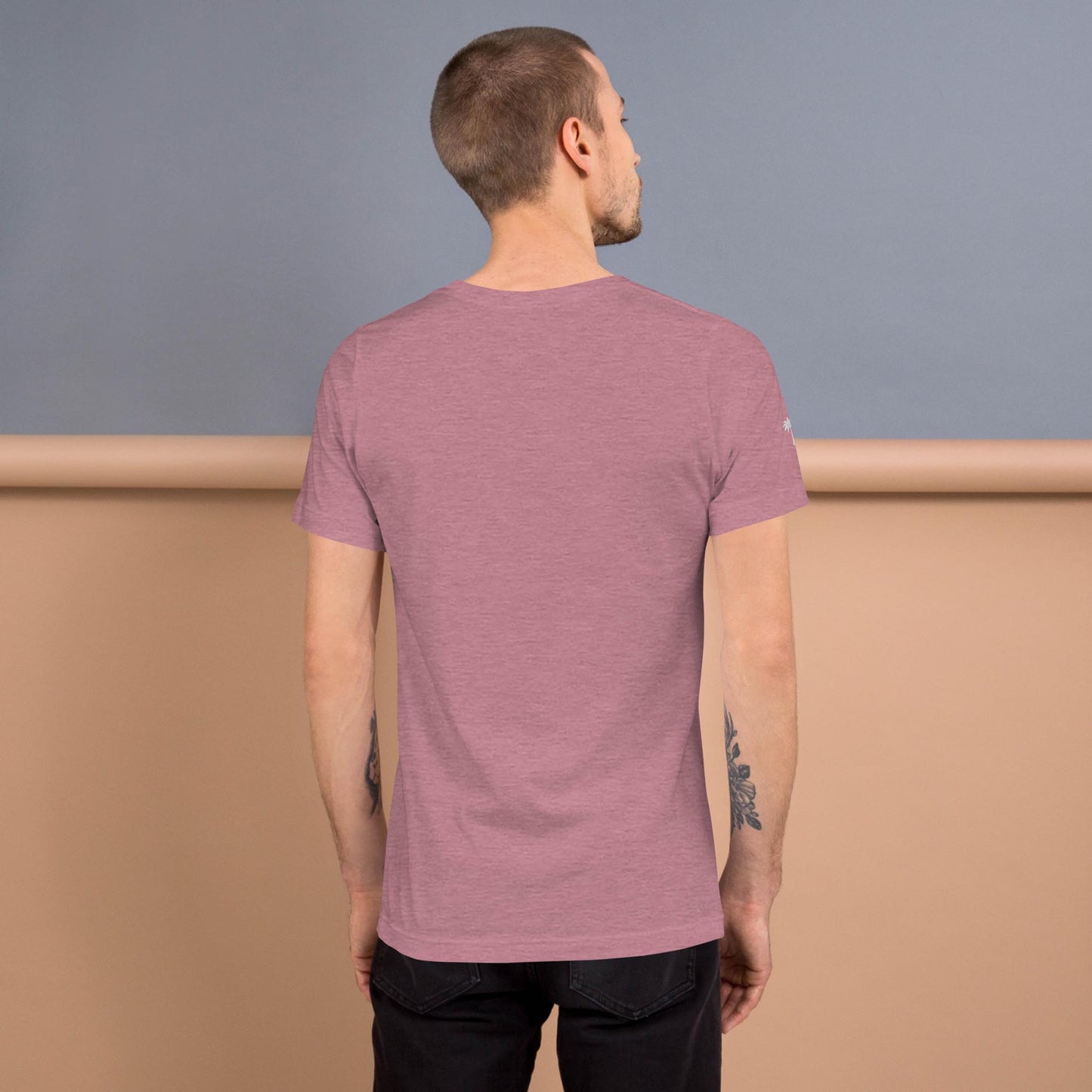 A man with short hair stands facing away, revealing the back of a mauve Mambí short-sleeve t-shirt. Tattoos cover both forearms, and he pairs the shirt with black pants. The background features beige and gray tones.