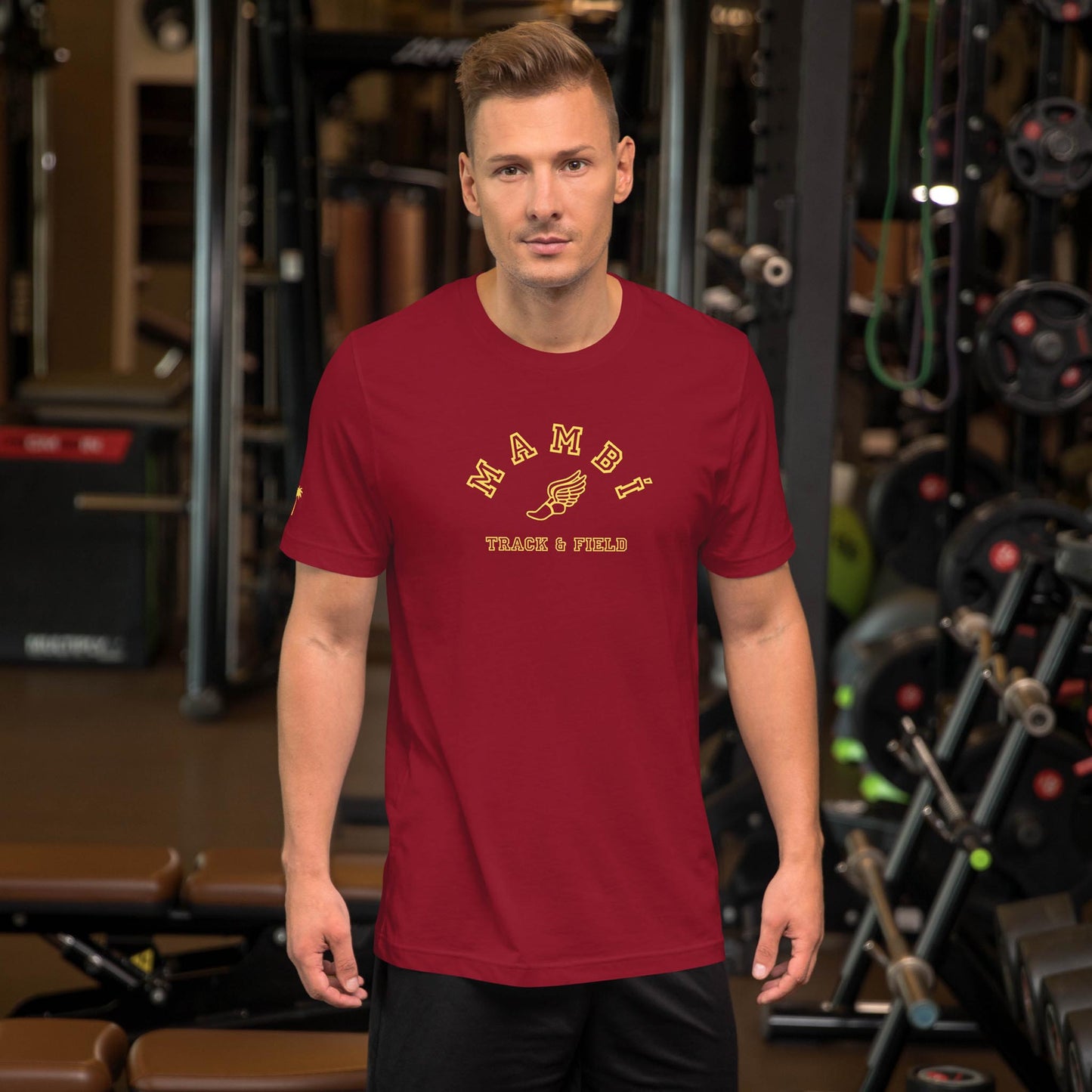 A man with short blonde hair stands in a gym, sporting a red Mambí Track & Field t-shirt and black shorts. Exercise equipment and weights are visible in the background.